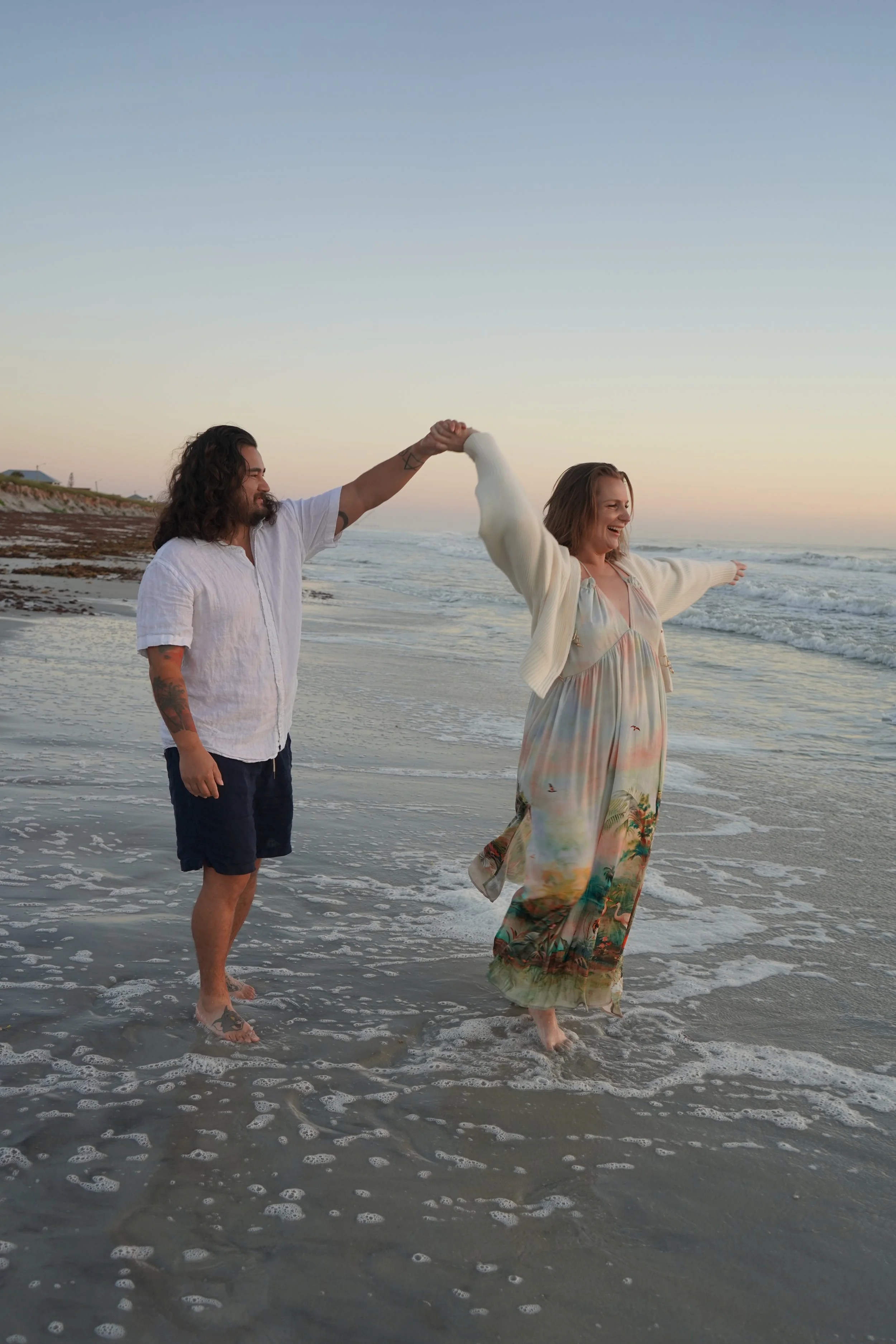 A woman and a man at a beach engagement, with the woman's arm raised and holding a sheer veil, and the man holding a bouquet of white baby's breath flowers in front of his face, standing against a rustic wooden wall.
