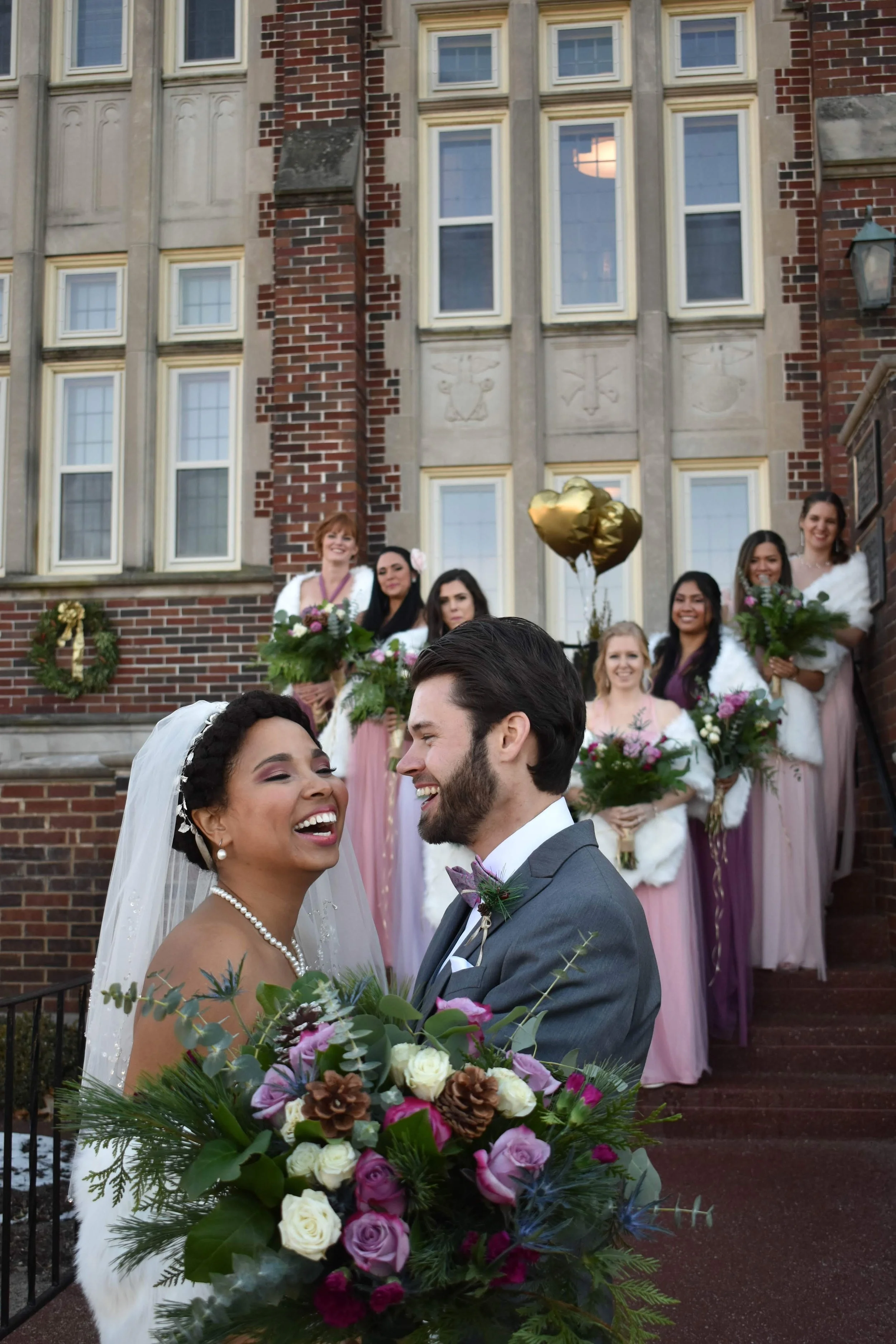 A couple dressed in wedding attire smiling and embracing outdoors, with the man in a black suit and the woman in a white lace wedding dress, wearing a floral crown.
