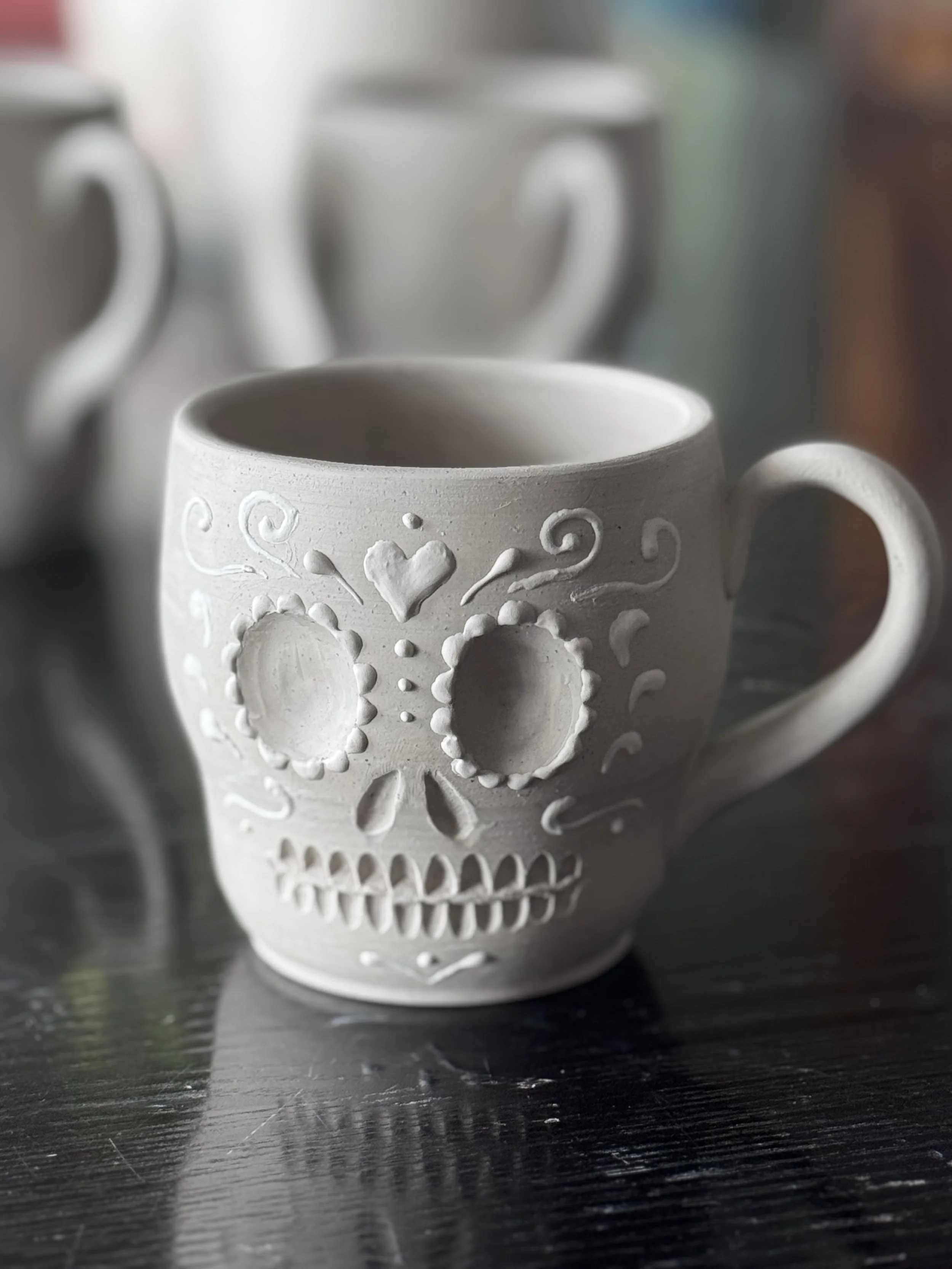 White ceramic mug with a carved sugar skull design, featuring carved eye and nose holes, teeth, and decorative patterns, placed on a dark wooden surface with other similar mugs in the background.