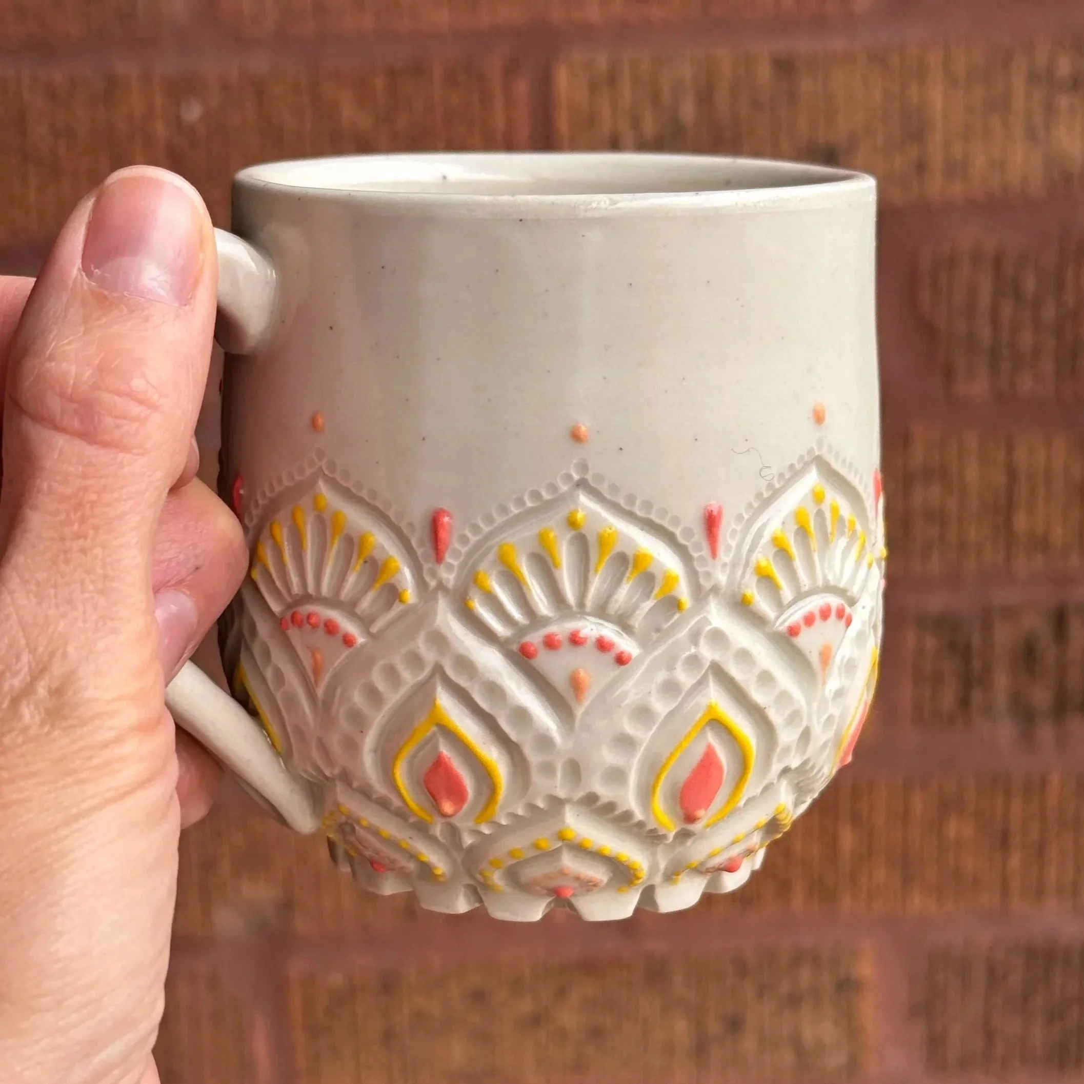 Close-up of a person's hand holding a carved white porcelain mug with pink, yellow, and peach slip dot decorations, against a brick background.