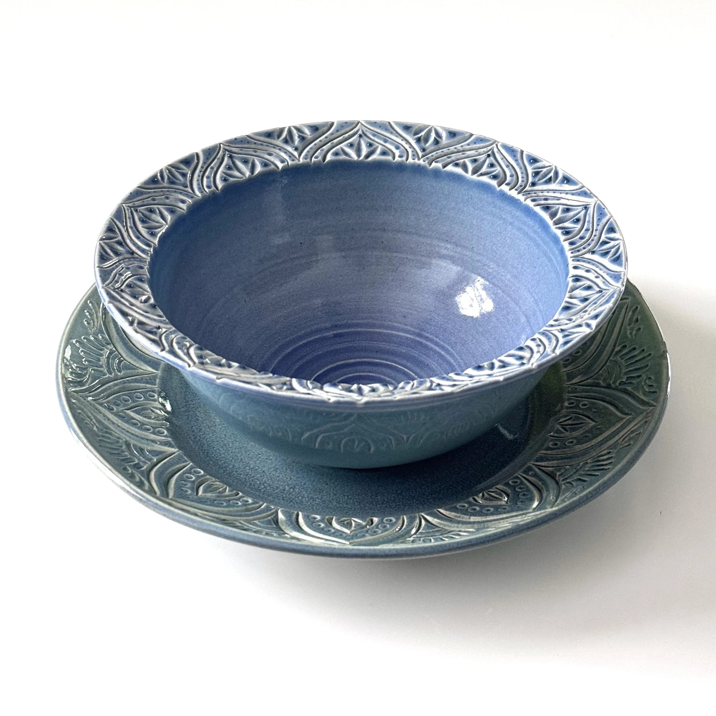 Handmade blue bowl with intricately carved rim set on a similarly carved serving plate on a white background