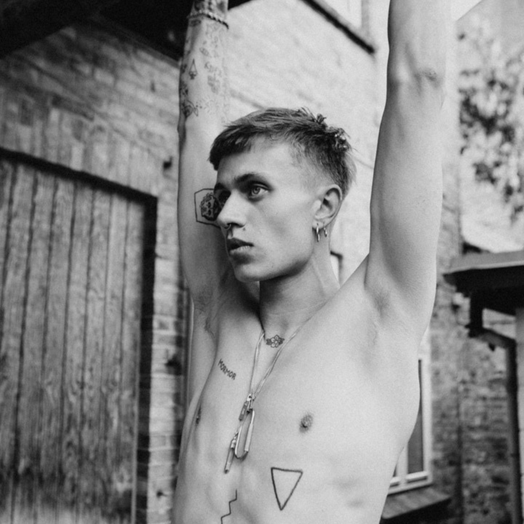 Black and white photo of a shirtless person with short hair, multiple tattoos, and jewelry, standing outdoors with arms raised against a brick building.