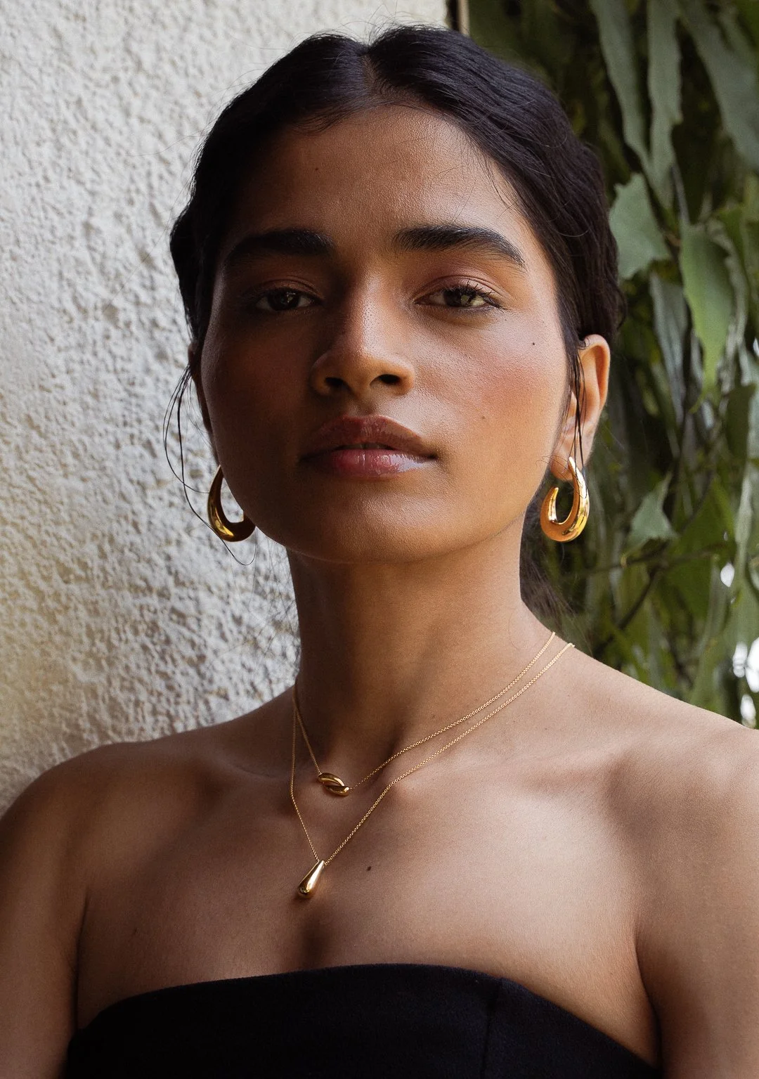 A woman with dark hair and medium skin tone wearing gold jewelry, including hoop earrings and layered necklaces, standing outdoors against a wall and greenery backdrop.