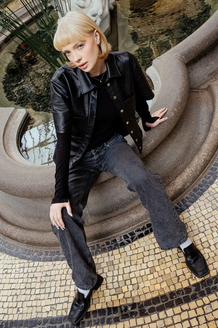 A young woman with blonde hair sitting on a stone bench next to a water fountain in a shopping mall or public indoor space, wearing a black leather jacket, black top, dark jeans, and black shoes.