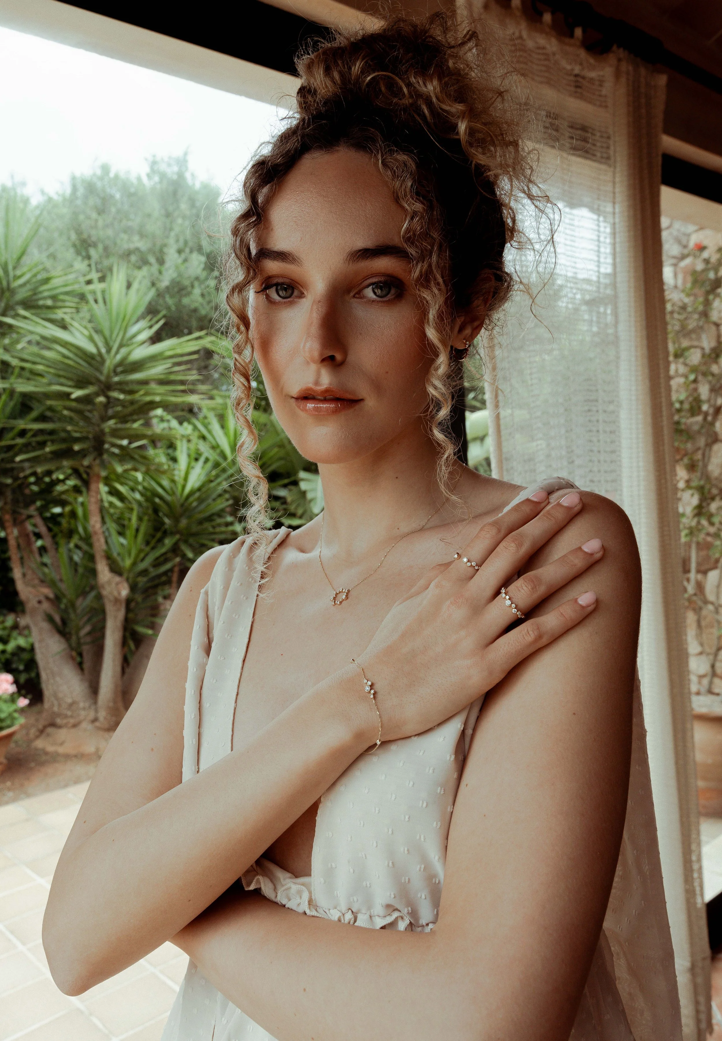 A young woman with curly hair and light skin posing indoors with a garden view visible through the window behind her. She is wearing a white dress and jewelry, including rings, a necklace, a bracelet, and earrings.