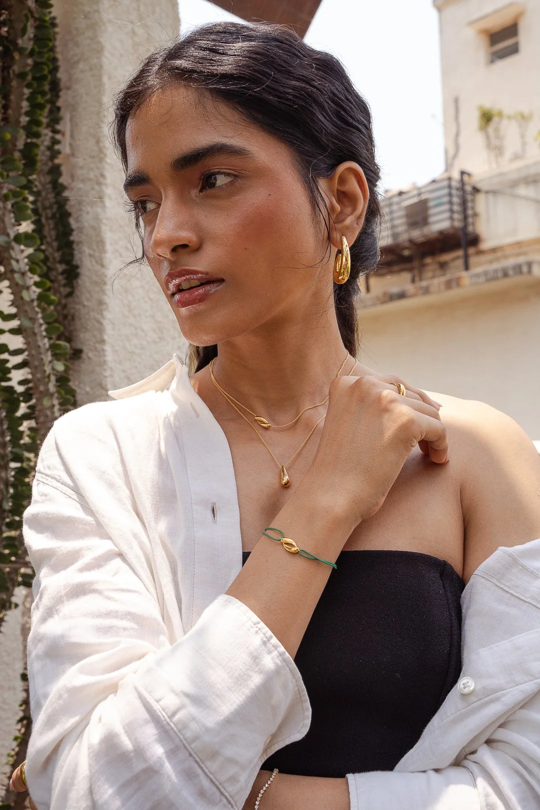 A woman with dark hair wearing gold jewelry, including earrings, necklaces, a bracelet, and rings, dressed in a black strapless top and white shirt, standing outdoors with buildings in the background.