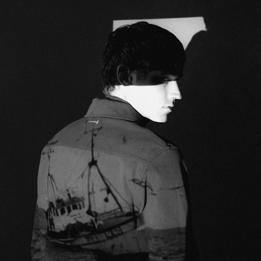 Black and white photo of a young man with dark, slicked-back hair, looking over his shoulder with a serious expression, illuminated by a high-contrast light, with a reflection of a boat or ship with masts and rigging projected onto his back.