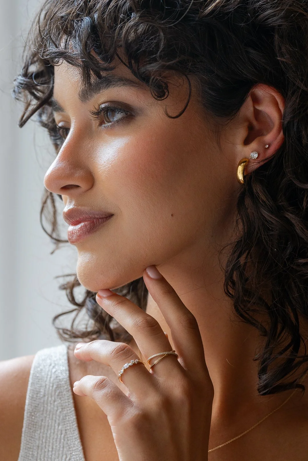 Close-up of a woman with short, curly hair, wearing gold and diamond earrings, and rings on her fingers, touching her face gently.