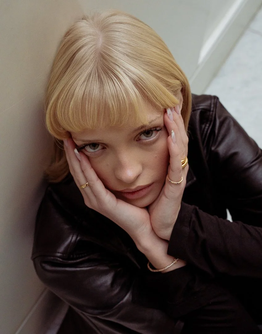 A young woman with blonde hair, wearing a dark brown leather jacket, sitting against a wall, resting her head in her hands, looking directly at the camera with a contemplative expression.