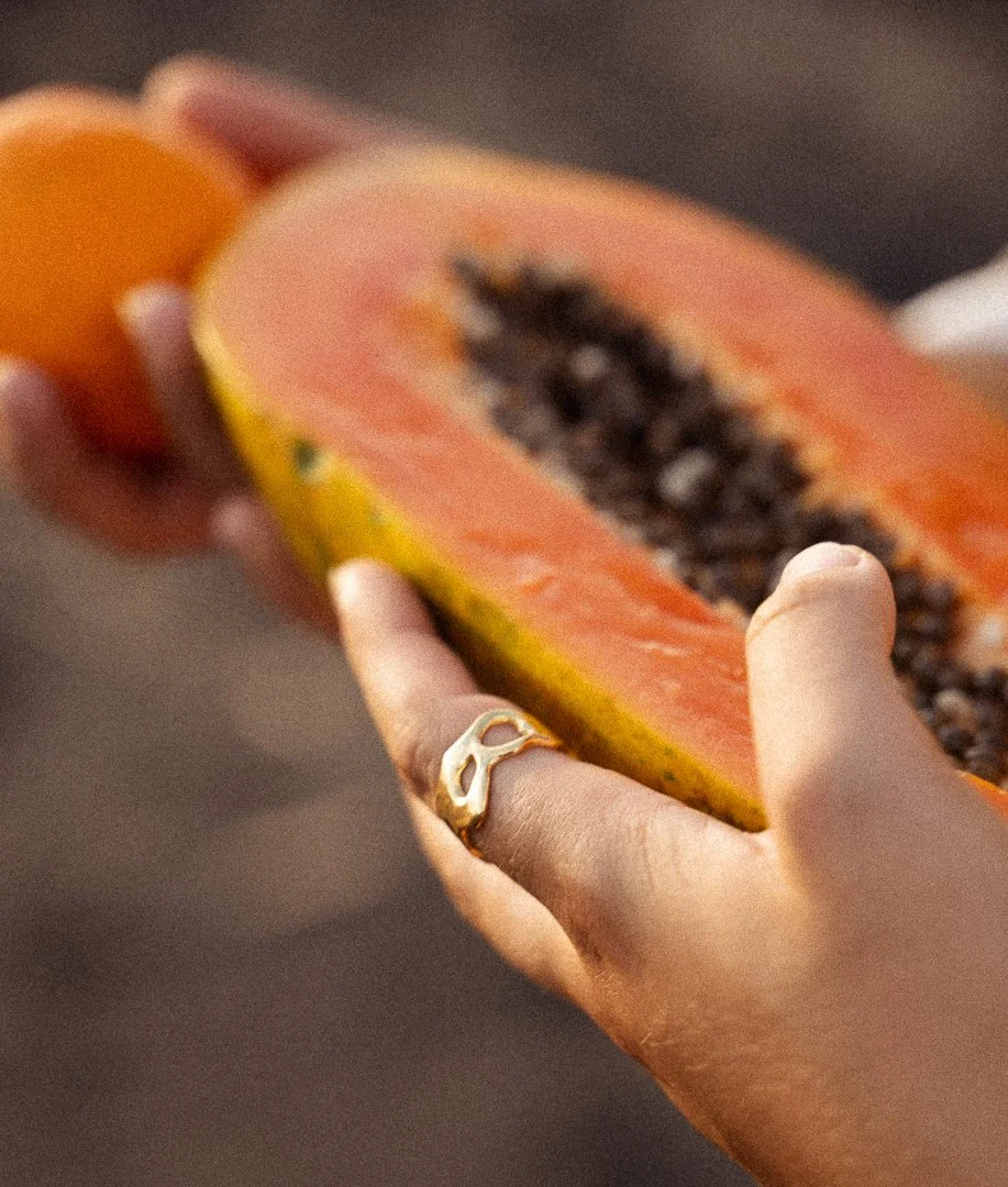 A person holding a halved papaya with orange flesh and black seeds, wearing a gold ring with a cutout design.