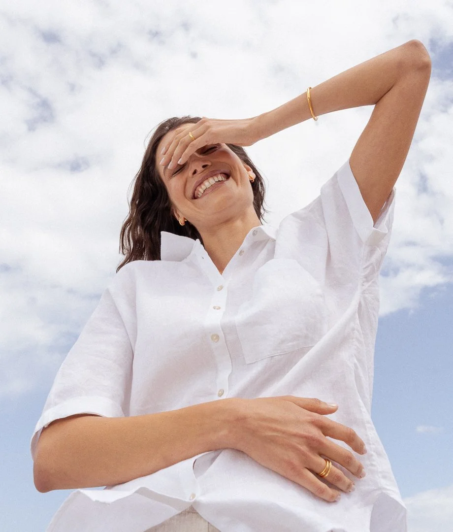 A woman smiling and laughing outdoors under a sky with scattered clouds, wearing a white shirt and gold jewelry, with her hand covering her eyes.