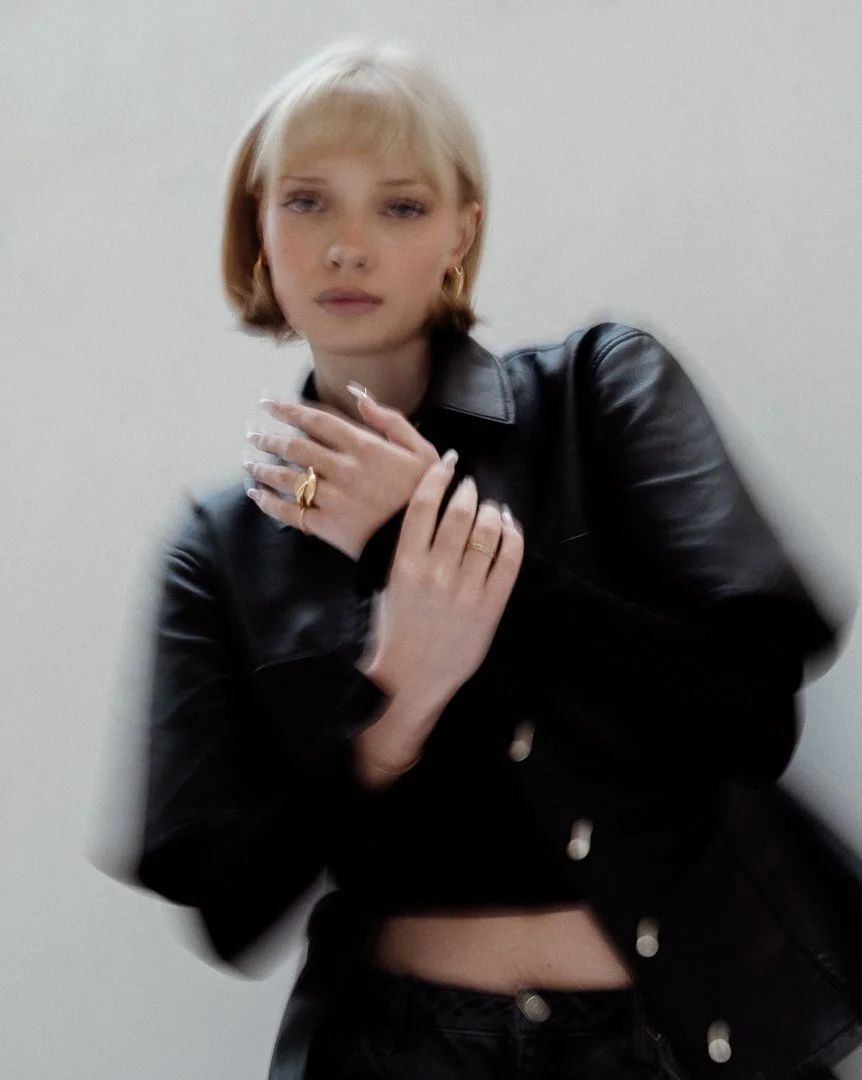 A young woman with blonde hair wearing a black leather jacket and jewelry, posing against a plain background.