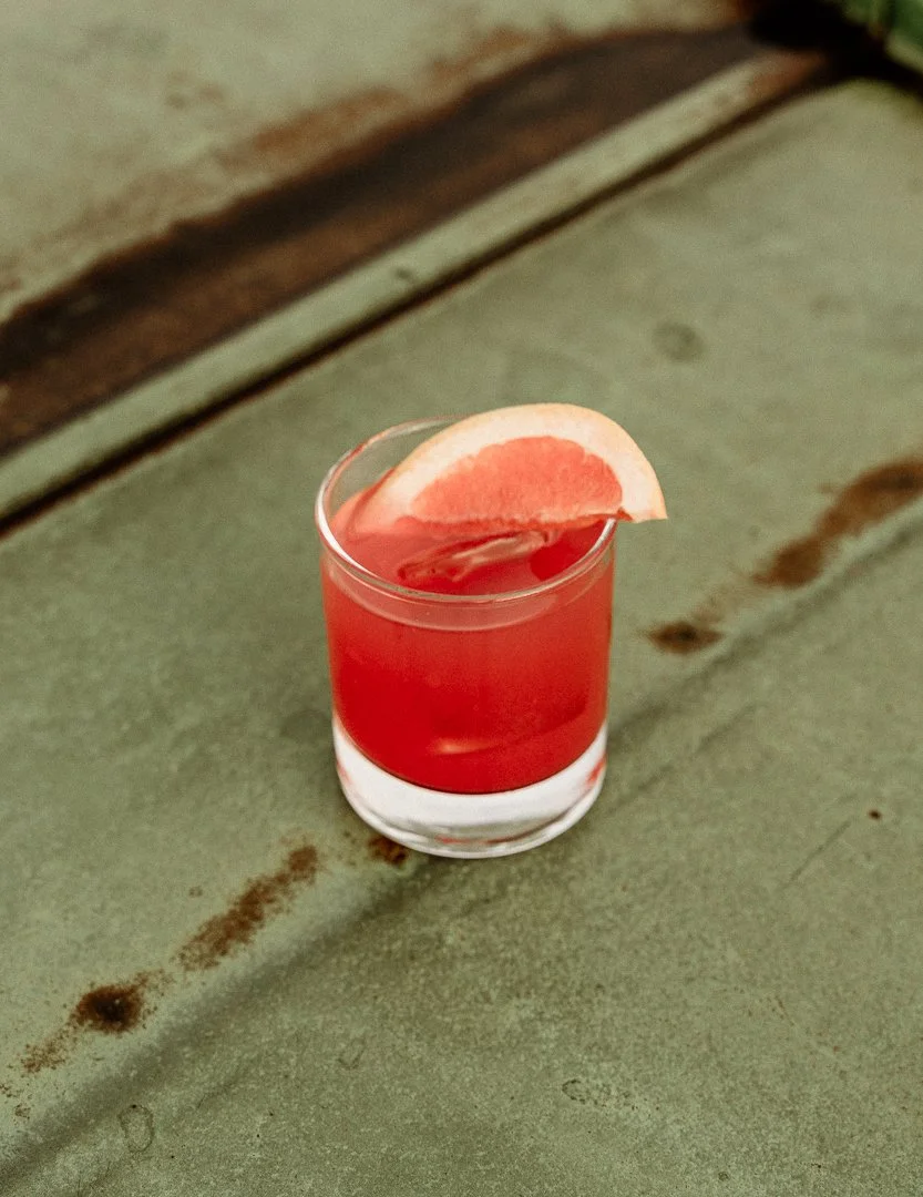 A clear glass with a red-colored drink garnished with a slice of grapefruit, placed on a rustic, green-painted surface.