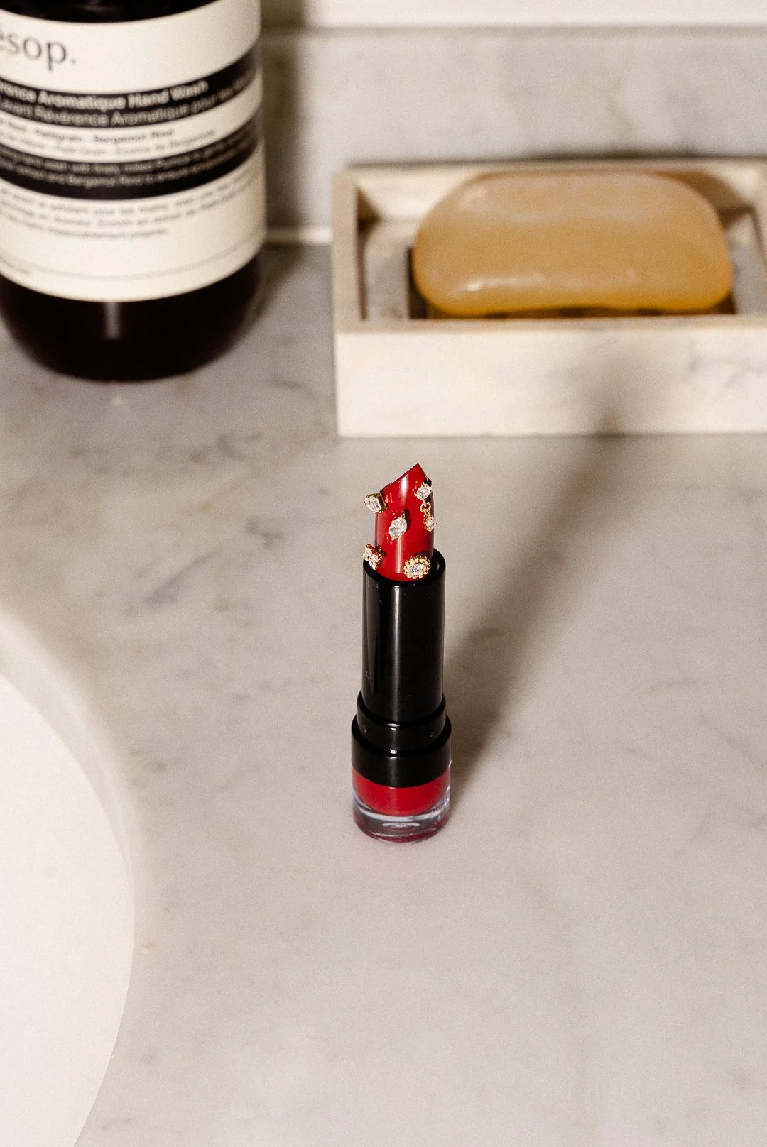 Red lipstick with diamond embellishments on a white marble surface, with a beige soap and a bottle of skincare product in the background.
