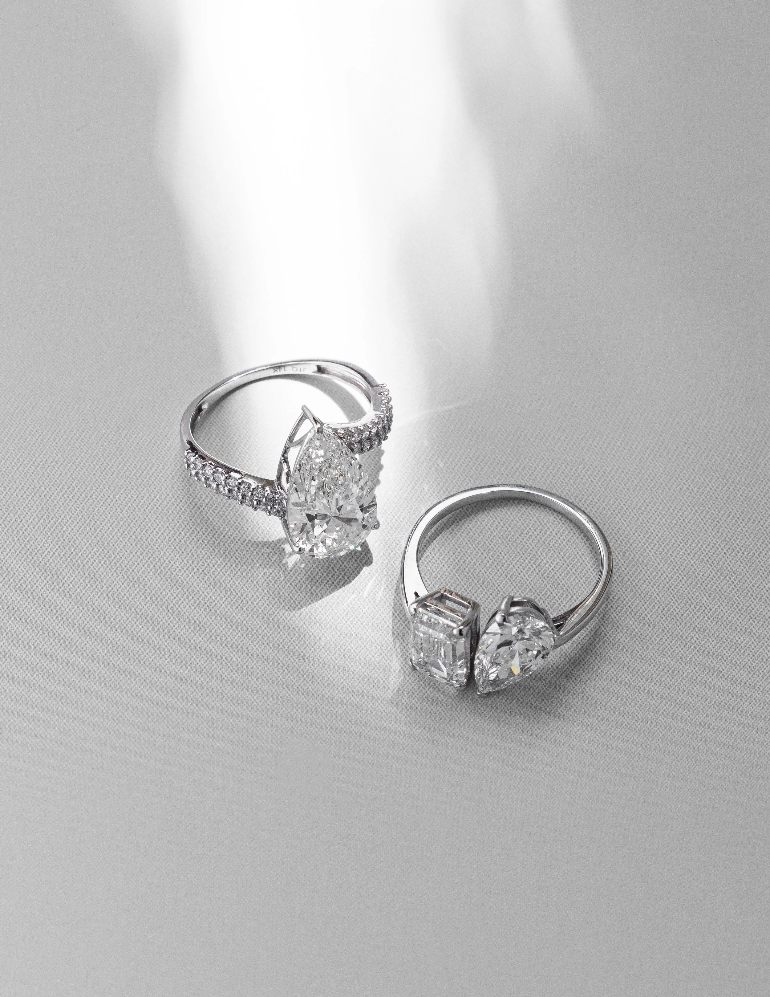 Two silver rings with large clear gemstones, one with a pear-shaped diamond and the other with a square and heart-shaped diamond, on a light gray surface.