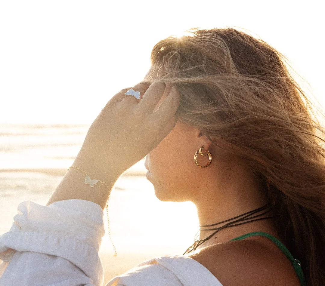 A woman with jewelry and a green strap top at the beach during sunset, with her hand touching her hair.