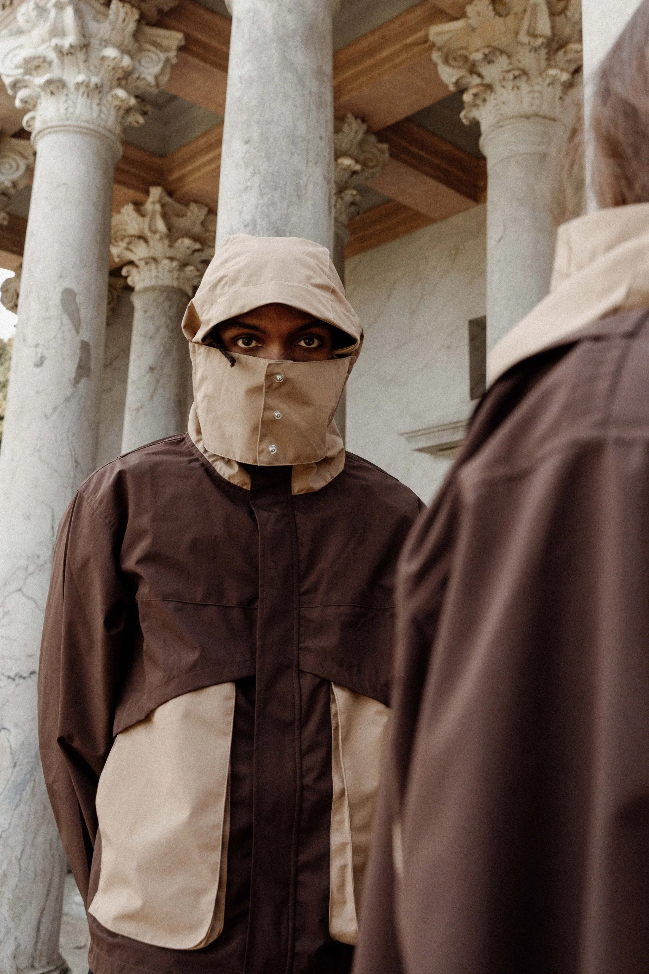 Person wearing a hood and face covering standing between marble columns, in front of a classical building.