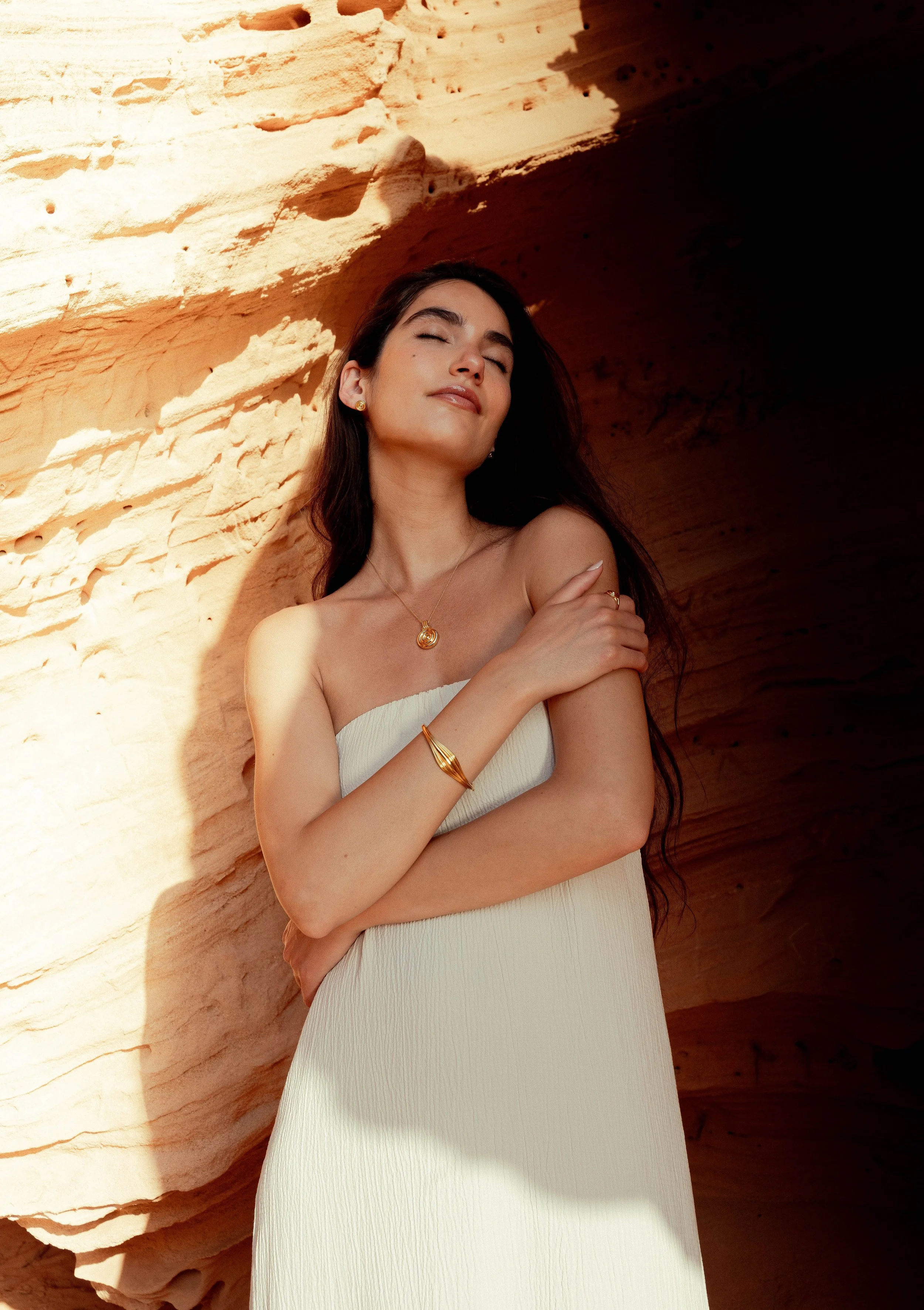 A young woman with long dark hair wearing jewelry and a strapless white dress, standing in front of a rock formation with sunlight casting shadows.