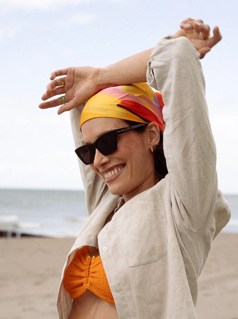 A woman smiling and enjoying herself on the beach, wearing sunglasses, a colorful headscarf, and a beige open shirt over an orange bikini top.