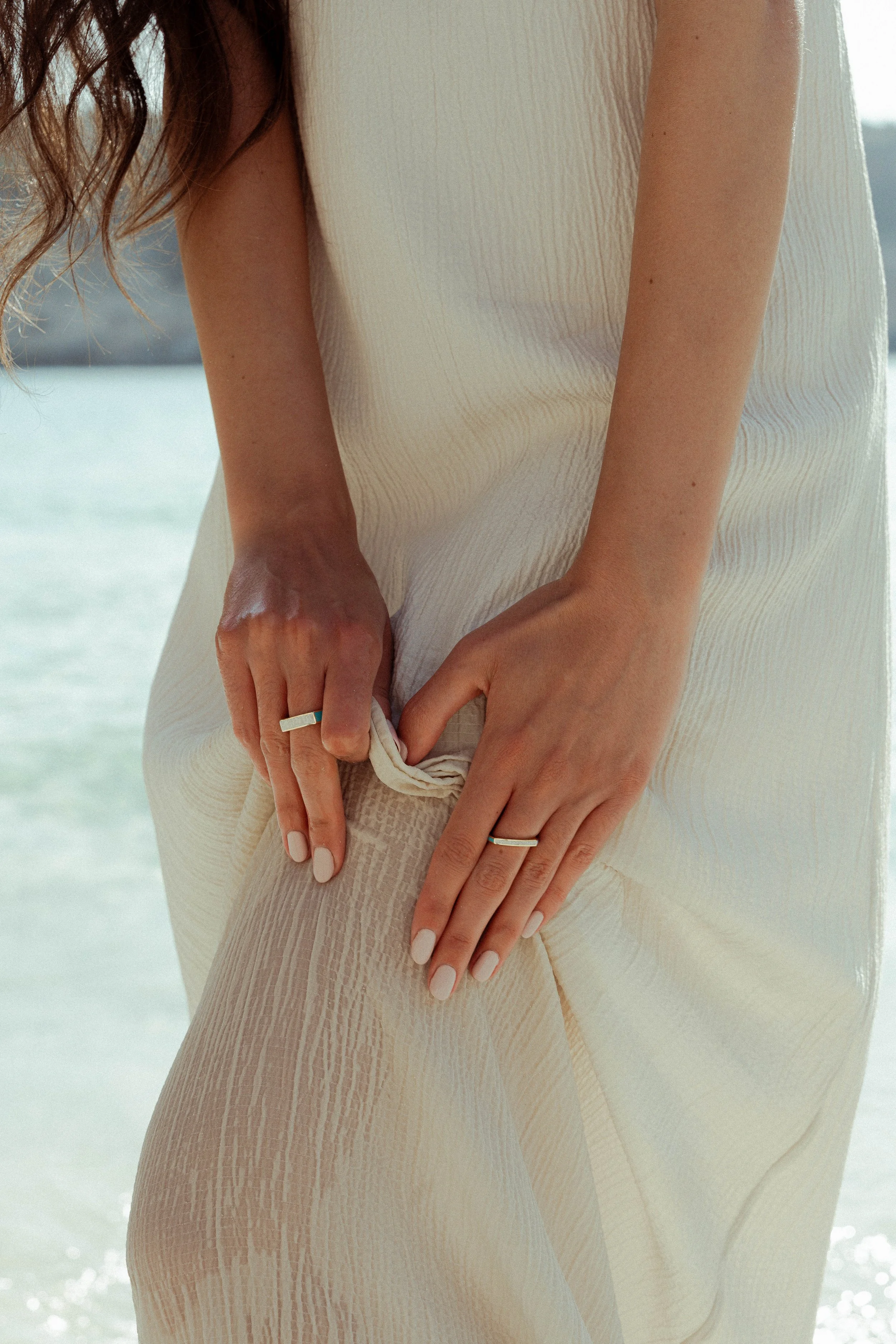 A person in a cream-colored outfit pulls their seam up, revealing tights, with both hands wearing matching rings. The background includes a body of water and distant shoreline, indicating a beach setting.