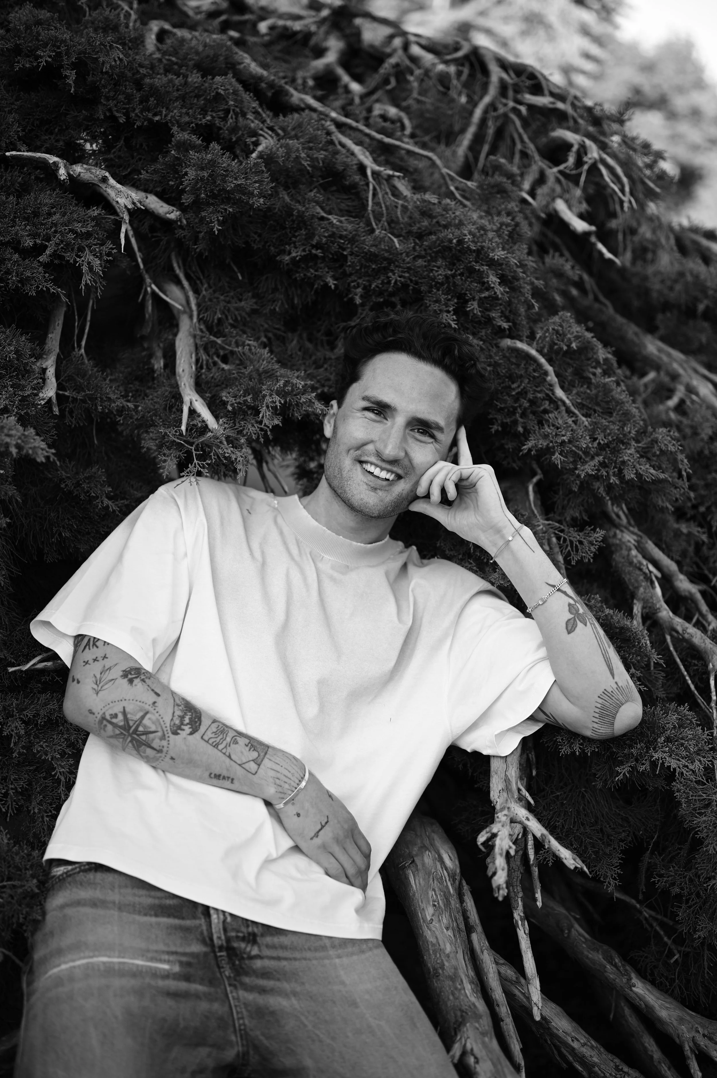 Black and white photo of a young man crouching outdoors, resting his head on his hand, wearing a chunky knit sweater.