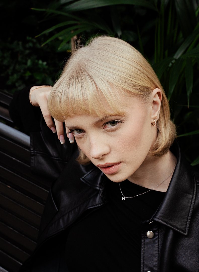 Close-up of a young woman with blonde hair, wearing a black leather jacket and jewelry, sitting on a bench with greenery in the background.