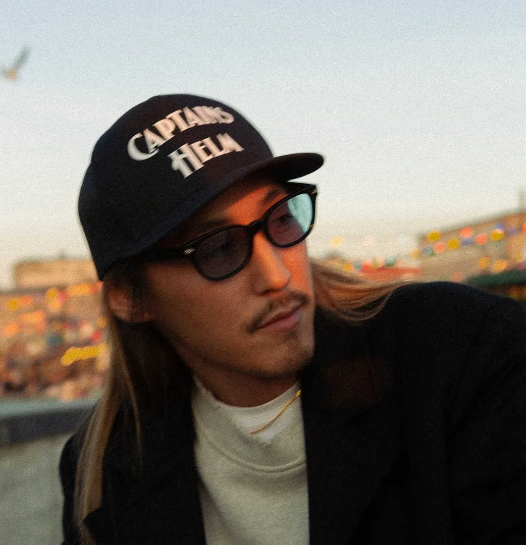 A young man with long hair, wearing sunglasses, a black baseball cap that says 'CAPTAINS OF THE FALL,' a white turtleneck, and a black jacket, outdoors with colorful lights in the background during dusk.