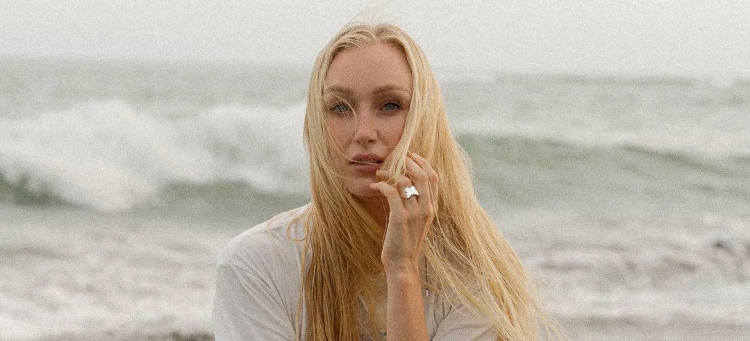 A young woman with long blonde hair and blue eyes standing on a beach with ocean waves in the background.
