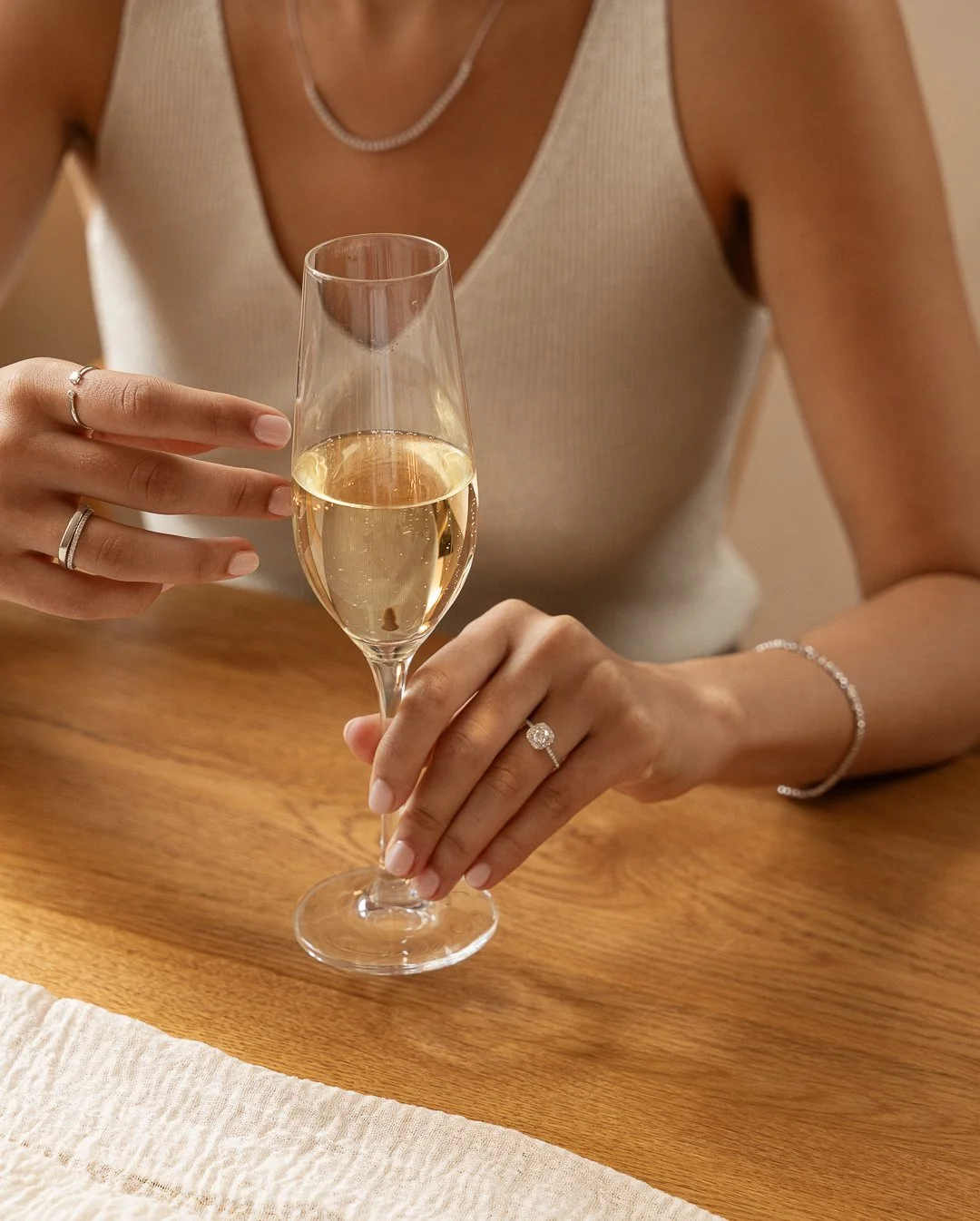 Woman in beige sleeveless top holding a glass of champagne, seated at a wooden table, wearing rings, bracelet, and necklace.