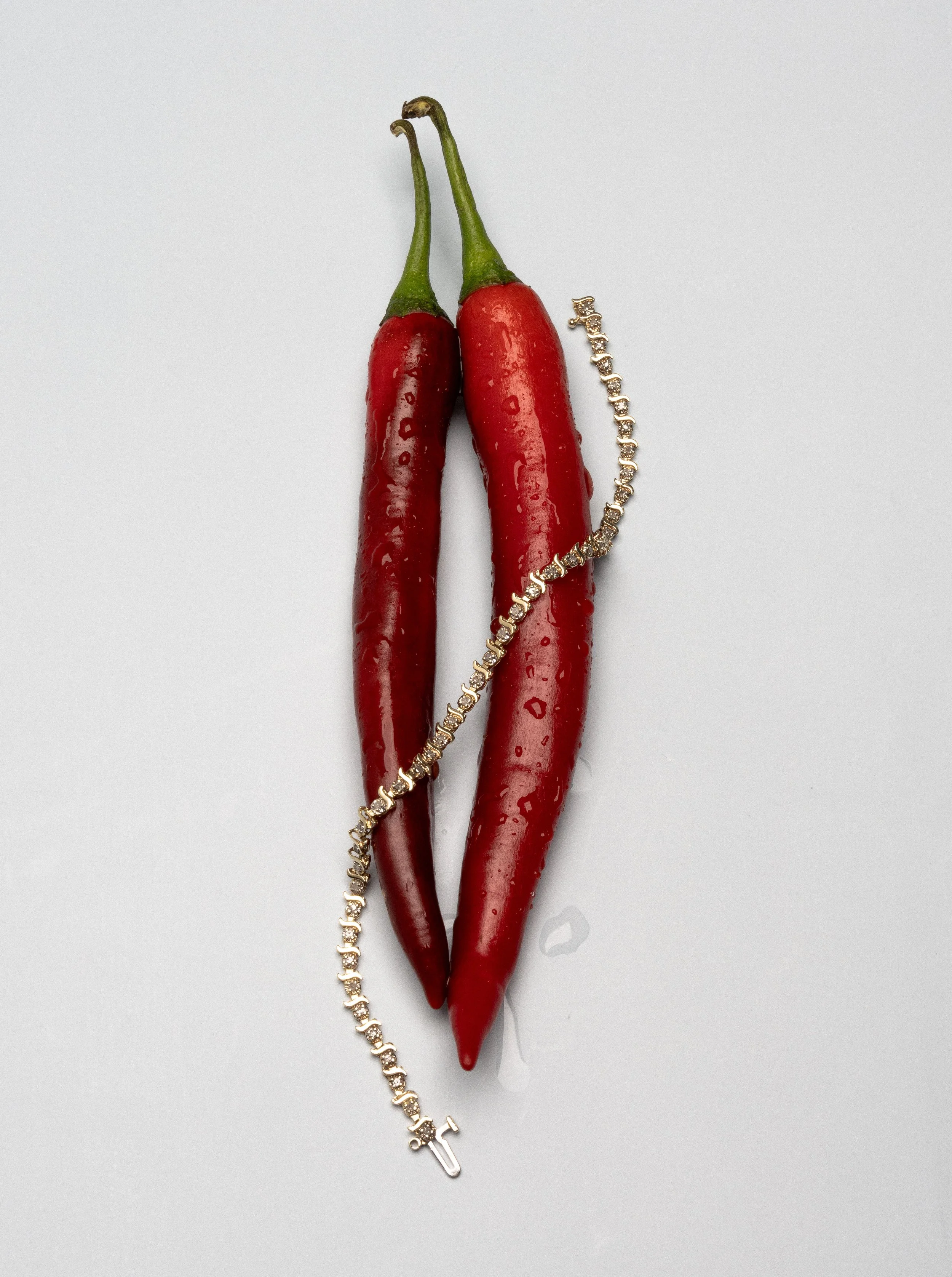 Two red chili peppers with a gold bracelet draped over them on a white background.