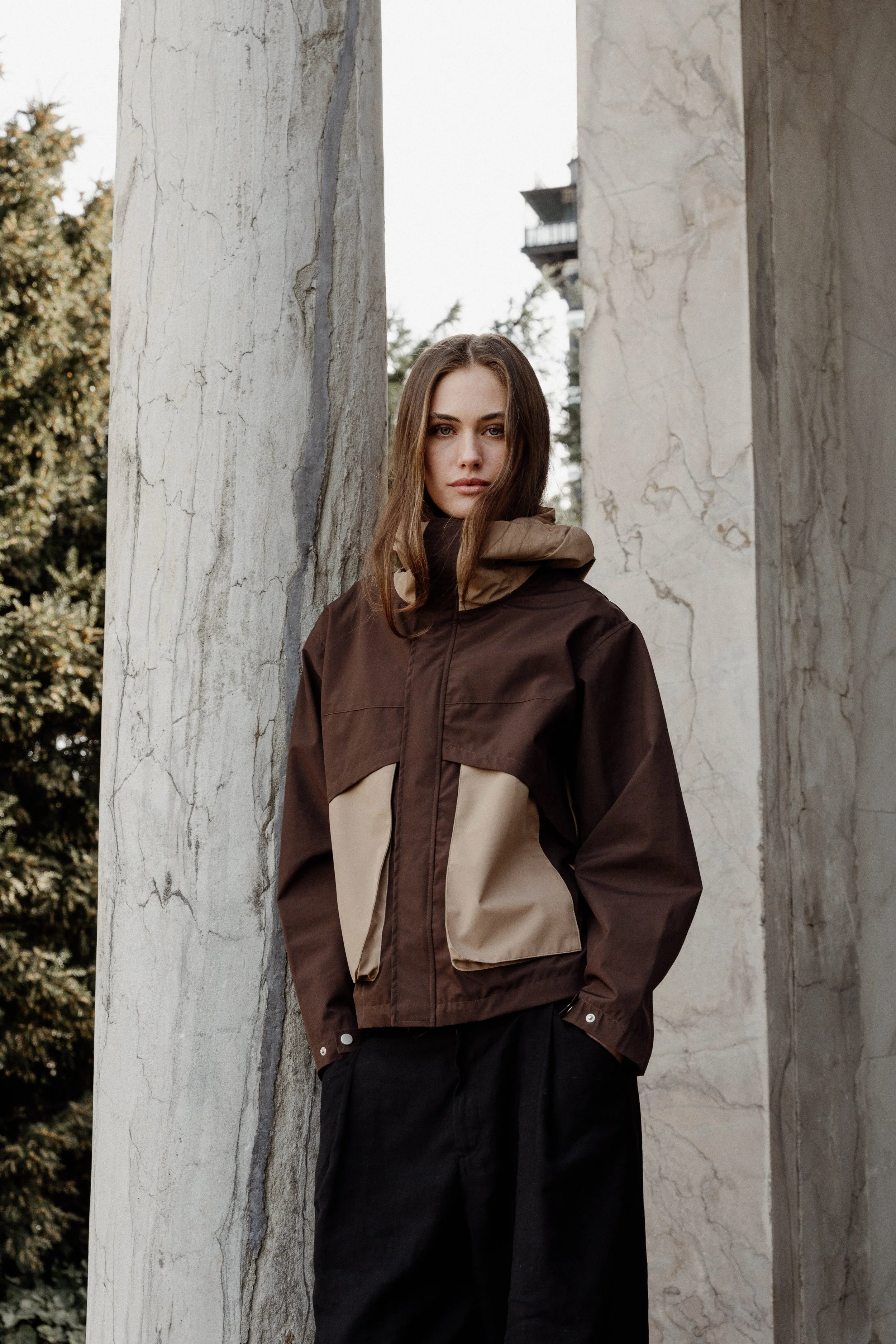 A young woman in a brown and beige jacket with a hood, standing outdoors against a large marble wall, with trees visible in the background.