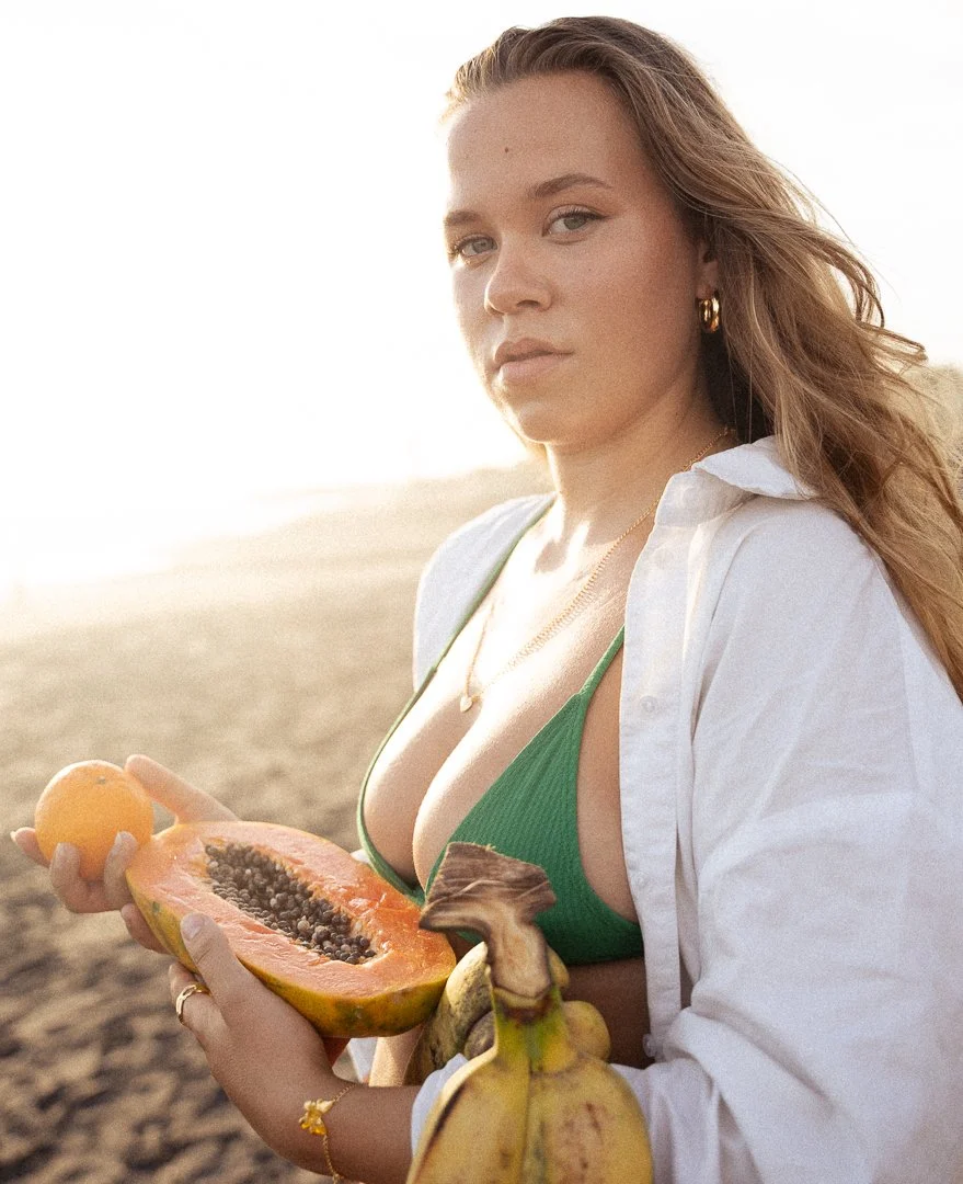 A woman on a beach holding a halved papaya, a banana, and a small orange, wearing a green bikini top and an open white shirt.