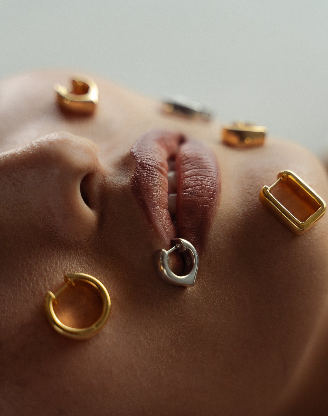 Close-up of a person's face with various gold and silver jewelry pieces on lips and cheeks, including hoops, rectangles, and a heart-shaped piece.