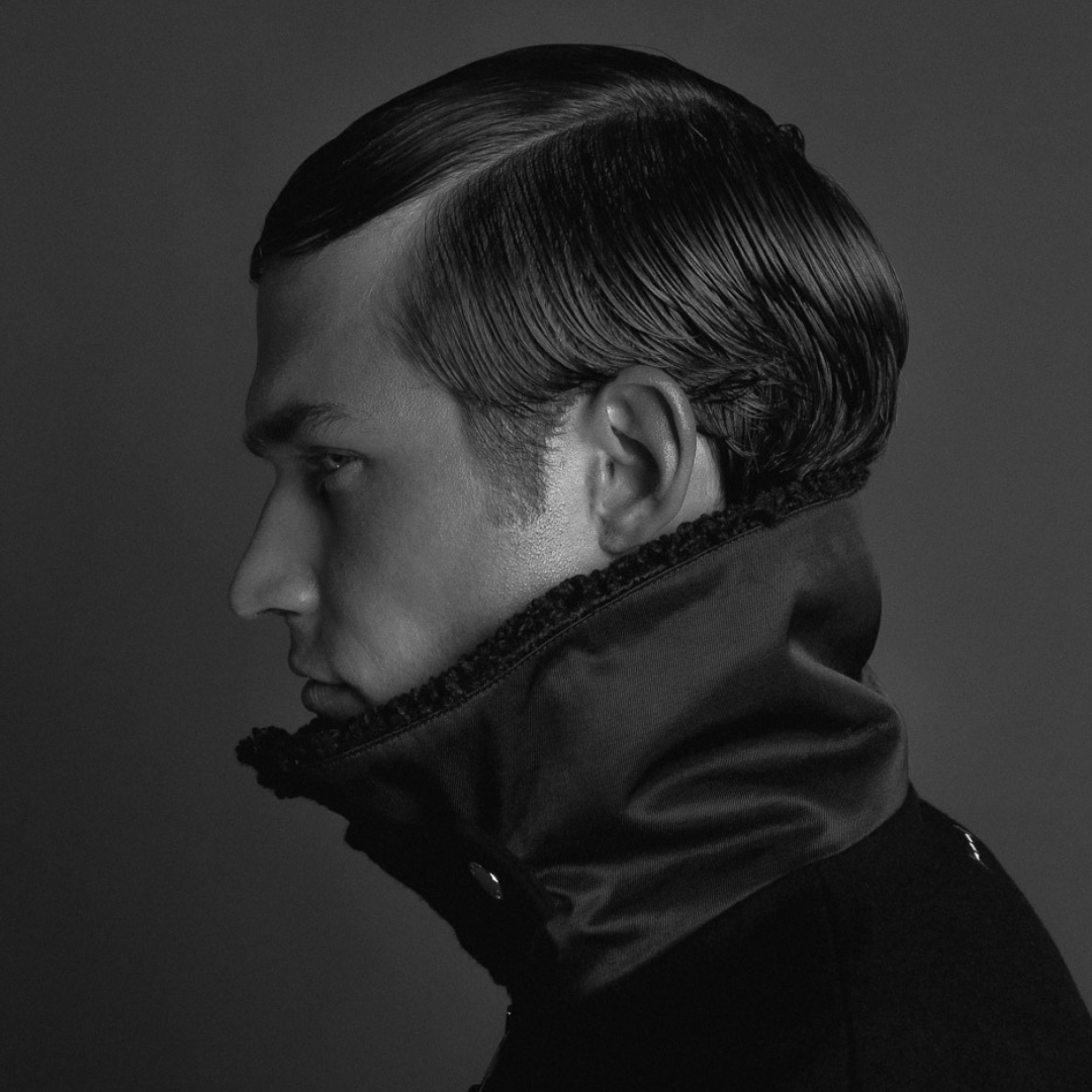 Black and white portrait of a young man with styled, slicked-back hair, wearing a high-collared jacket.