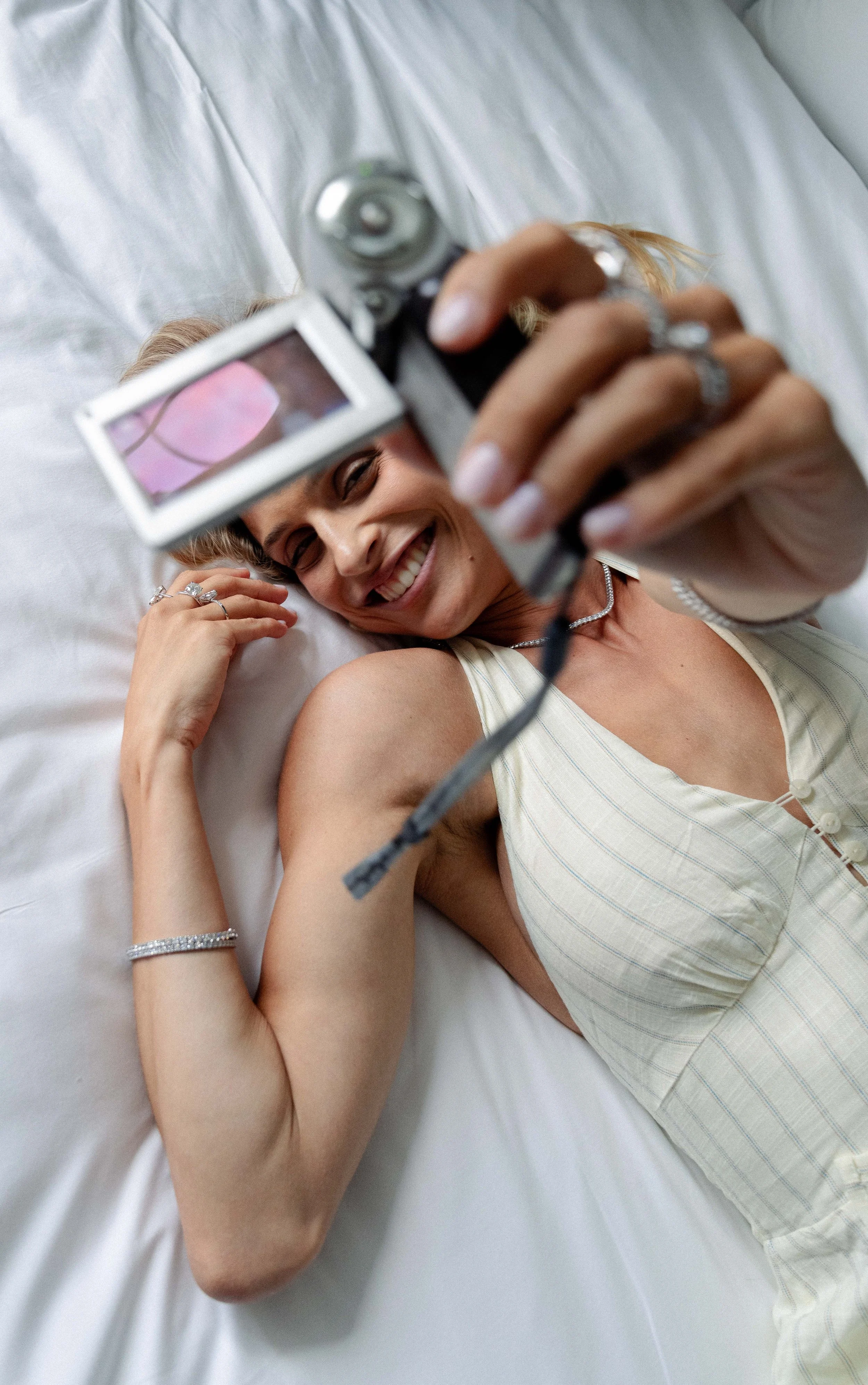 Woman smiling while lying on her bed, taking a selfie with a camera, wearing a light-colored sleeveless top and jewelry.