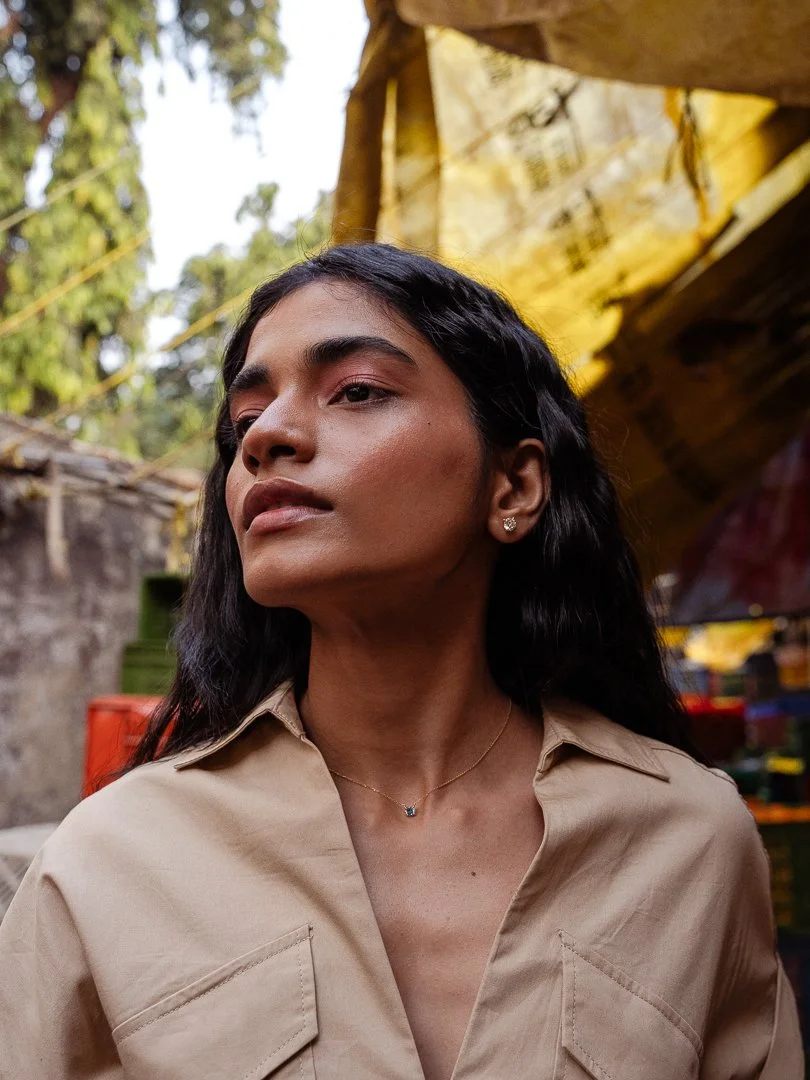 A young woman with long dark hair and tan skin standing outdoors, wearing a beige shirt, small earrings, and a delicate necklace, with blurry colorful items and trees in the background.