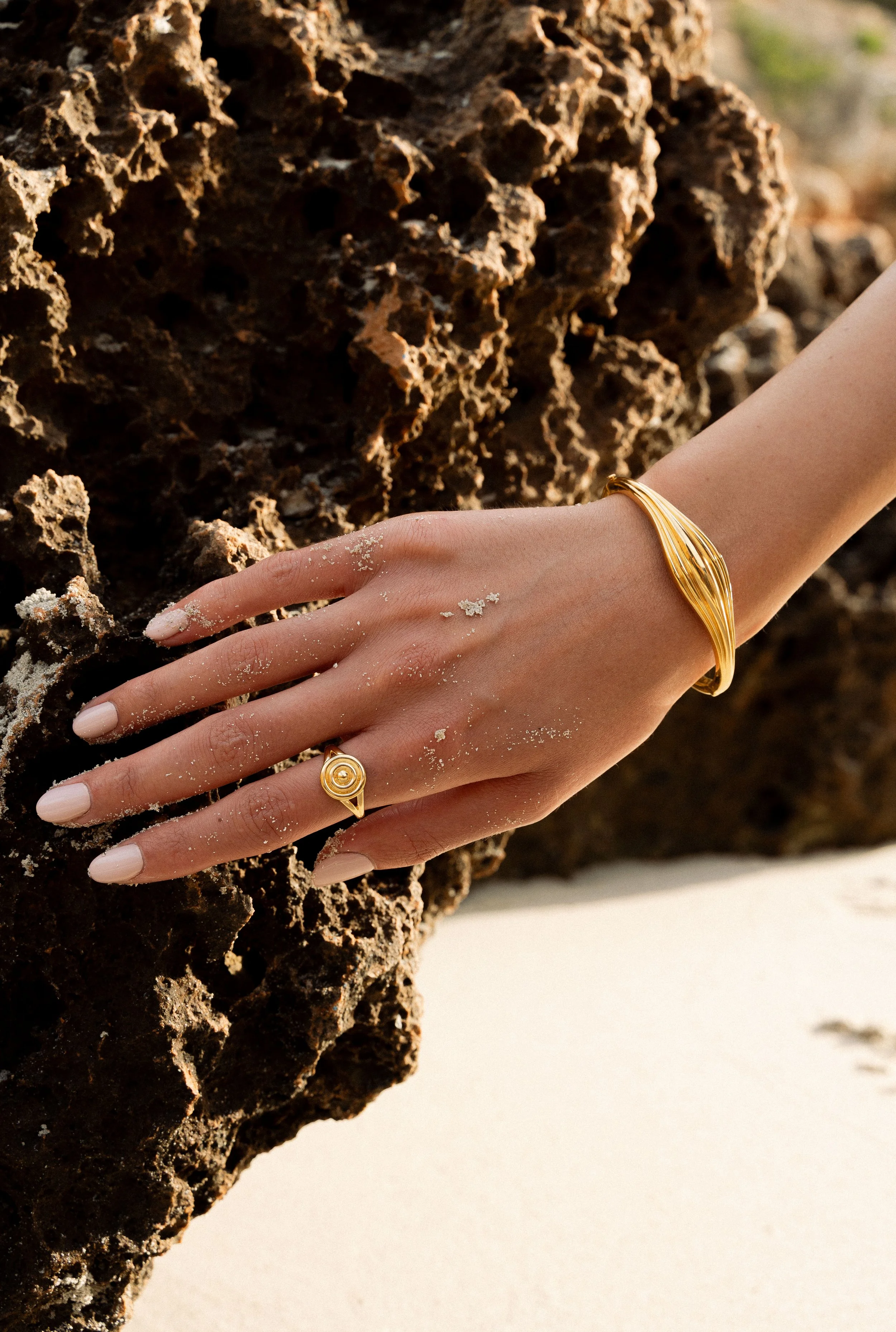 A woman's hand with a gold bracelet and ring, resting on a rocky surface with sand on her fingers and a sandy beach in the background.