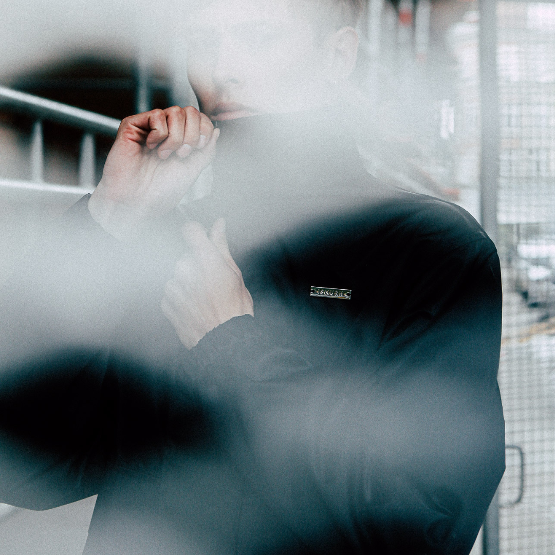 A person in a black jacket biting a sleeve on their right arm, with their face partially visible through a foggy or blurry overlay, standing outdoors near a metal railing and a wire fence.