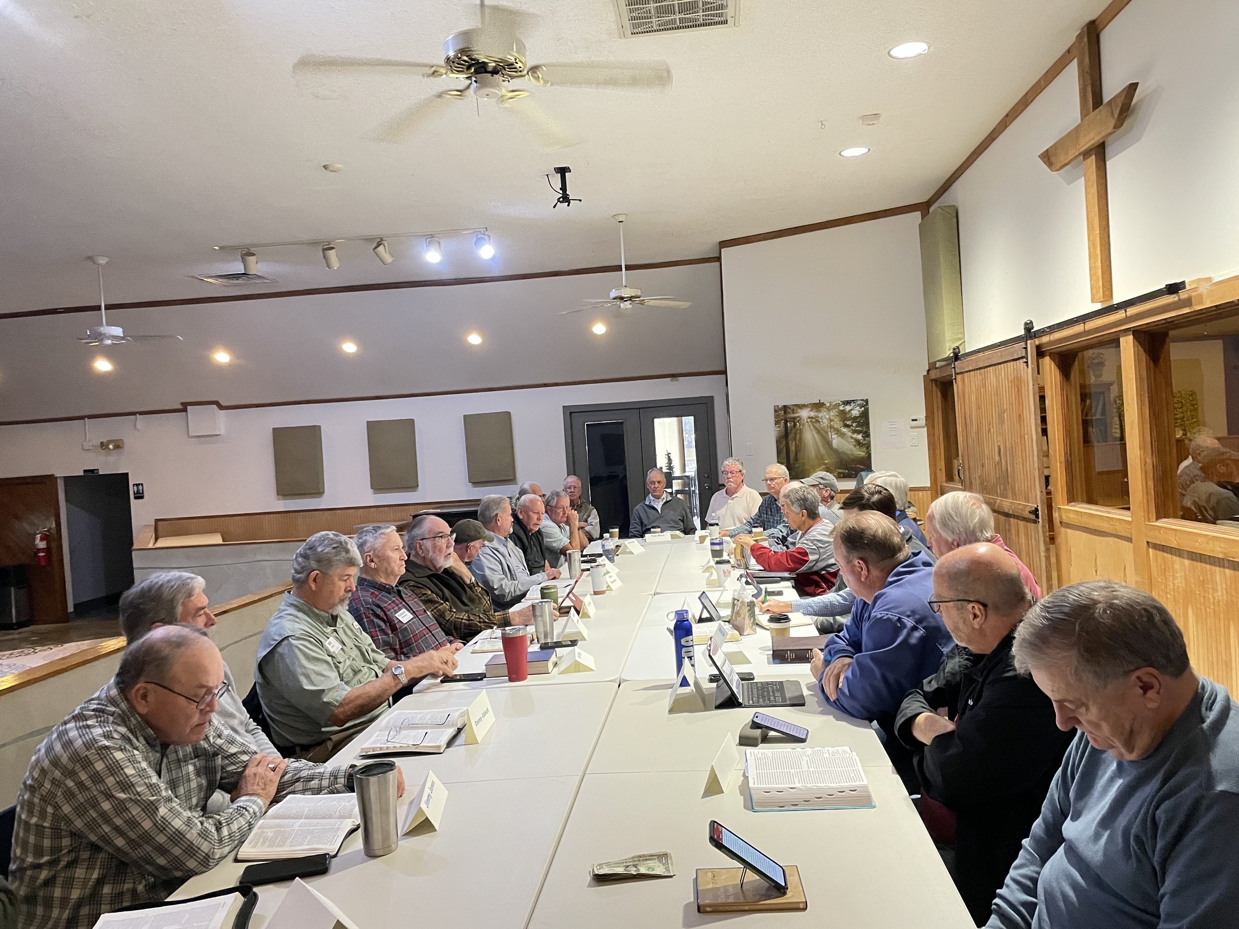A large group of elderly men and women seated around a long conference table in a meeting room. Several have open books or papers in front of them, and some are using laptops or smartphones.