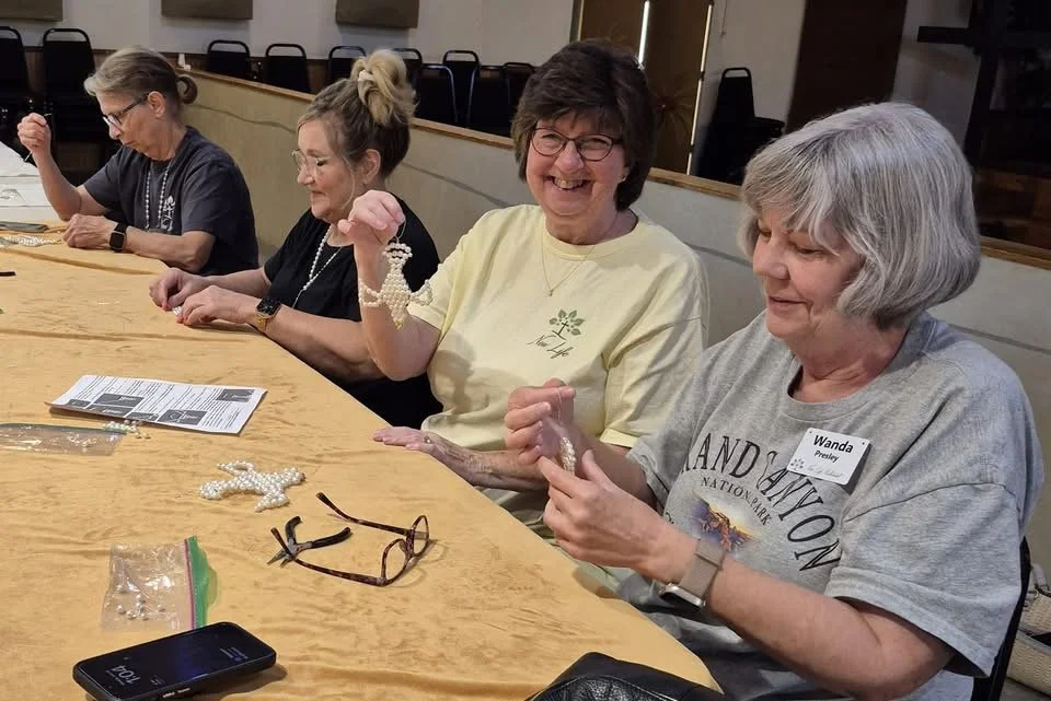 Group of women sitting at a table, working on craft projects with beads and jewelry-making supplies, one woman smiling and holding beads, another wearing a name tag that says 'Wanda.'