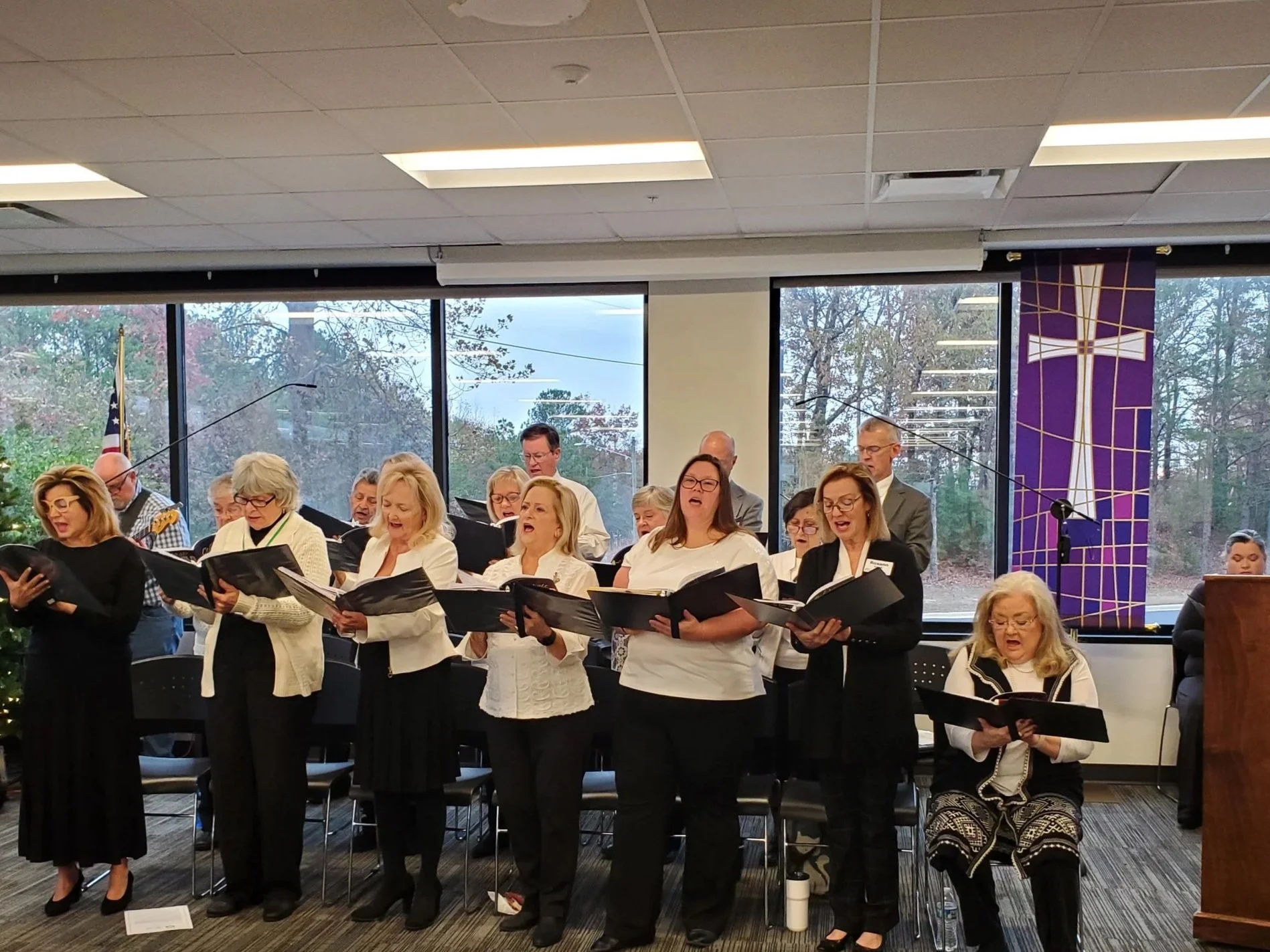 A choir of men and women singing during a church service or event in a room with large windows, some holding black folders, with Christmas decorations and purple church banners.