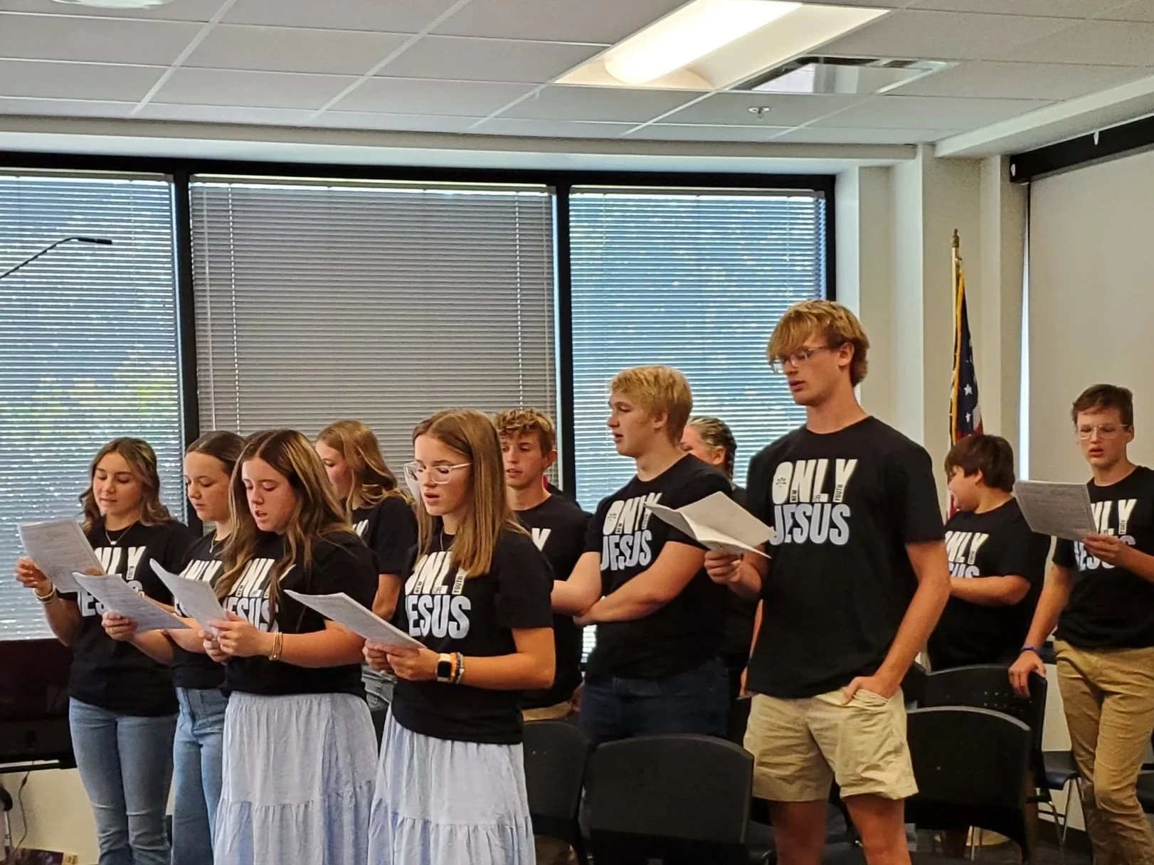 Group of teenagers standing in a classroom, singing or reading from papers, wearing matching black T-shirts that say 'ONLY JESUS'.