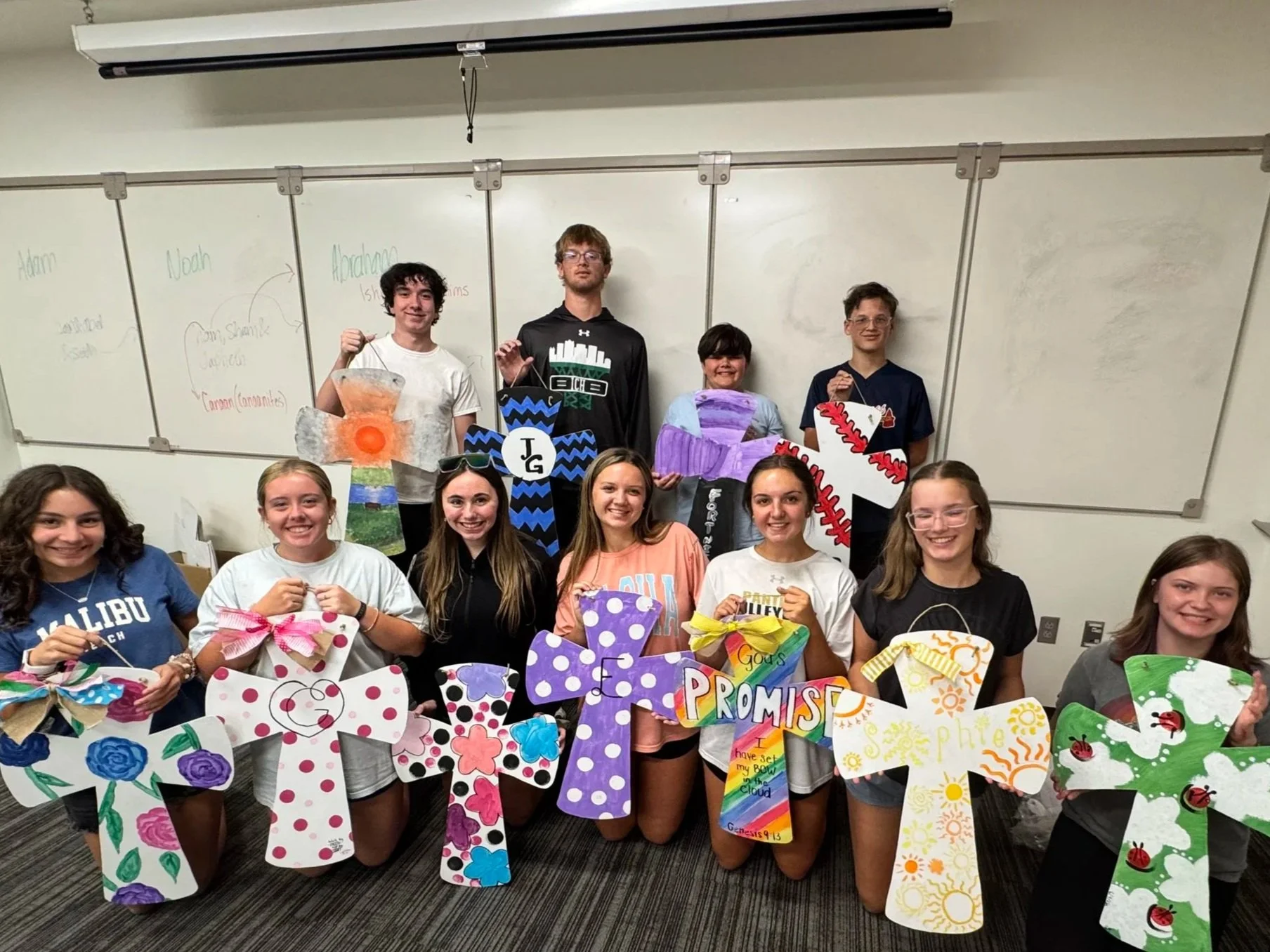 Group of ten students in a classroom holding decorated large wooden crosses with various patterns and colors.