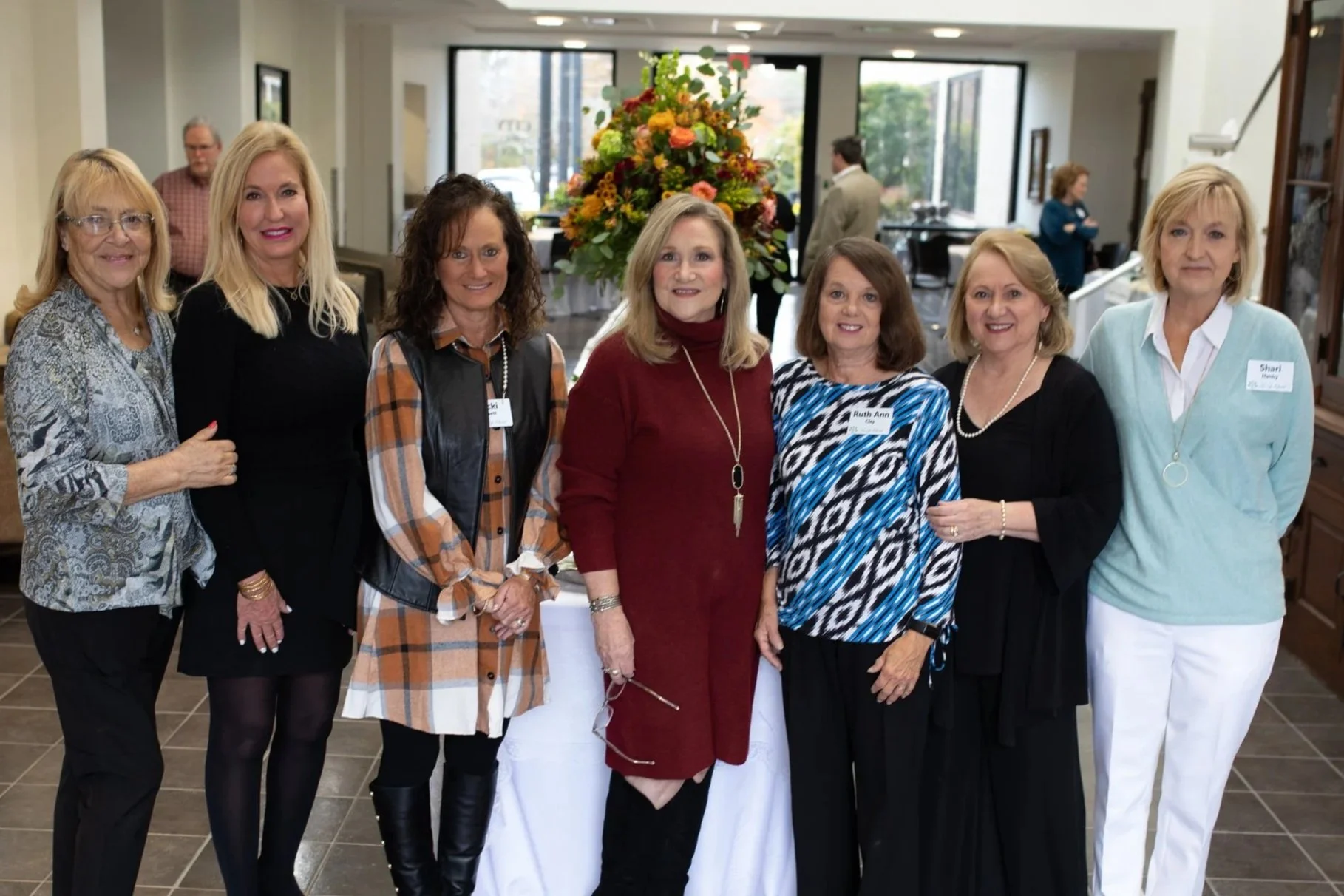 Group of seven women standing indoors in front of a large floral arrangement, smiling at the camera.
