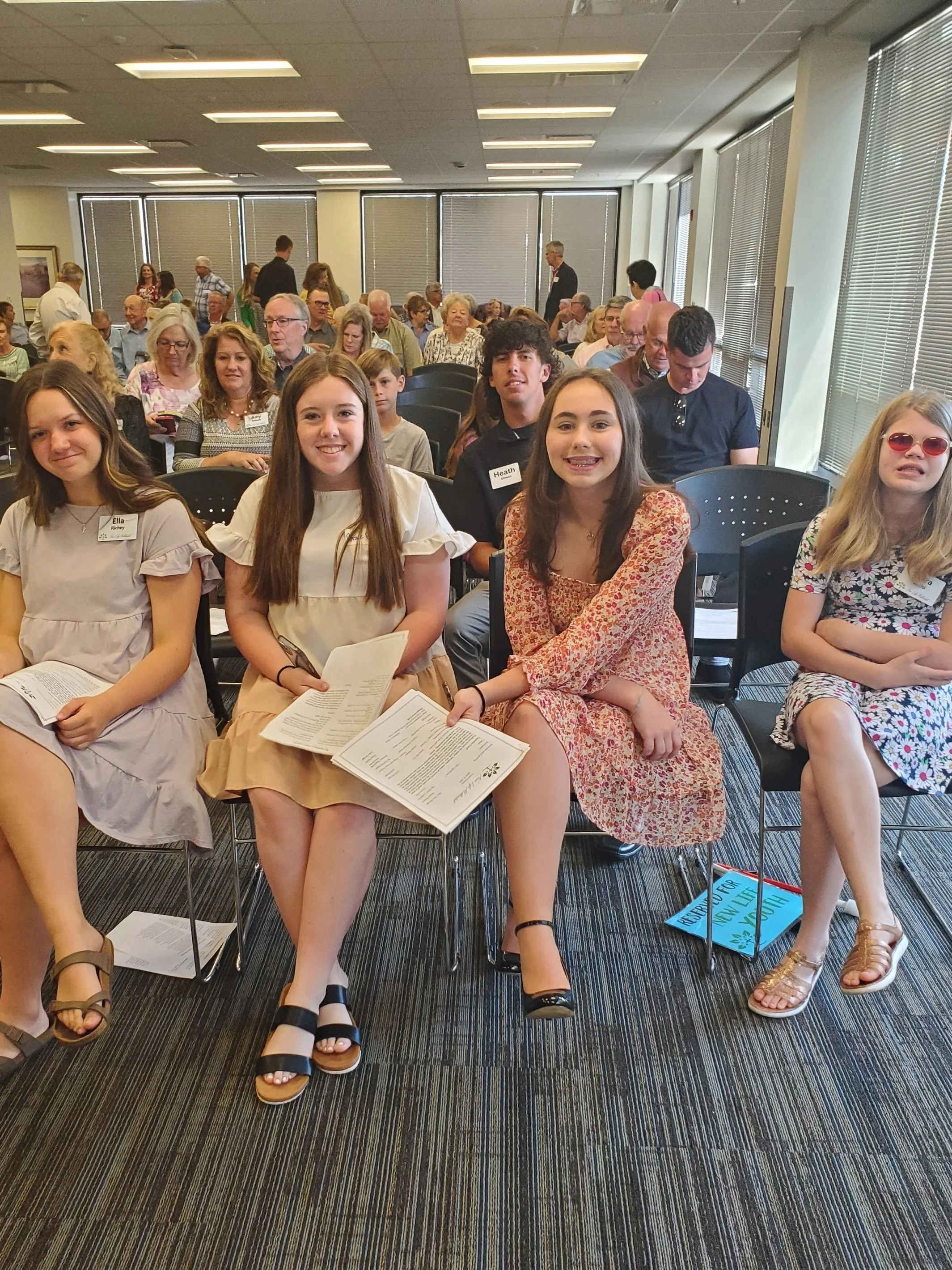 Group of young people, mostly girls, sitting in an indoor conference or event room, with many in the background, some holding papers, with large windows and blinds behind them.