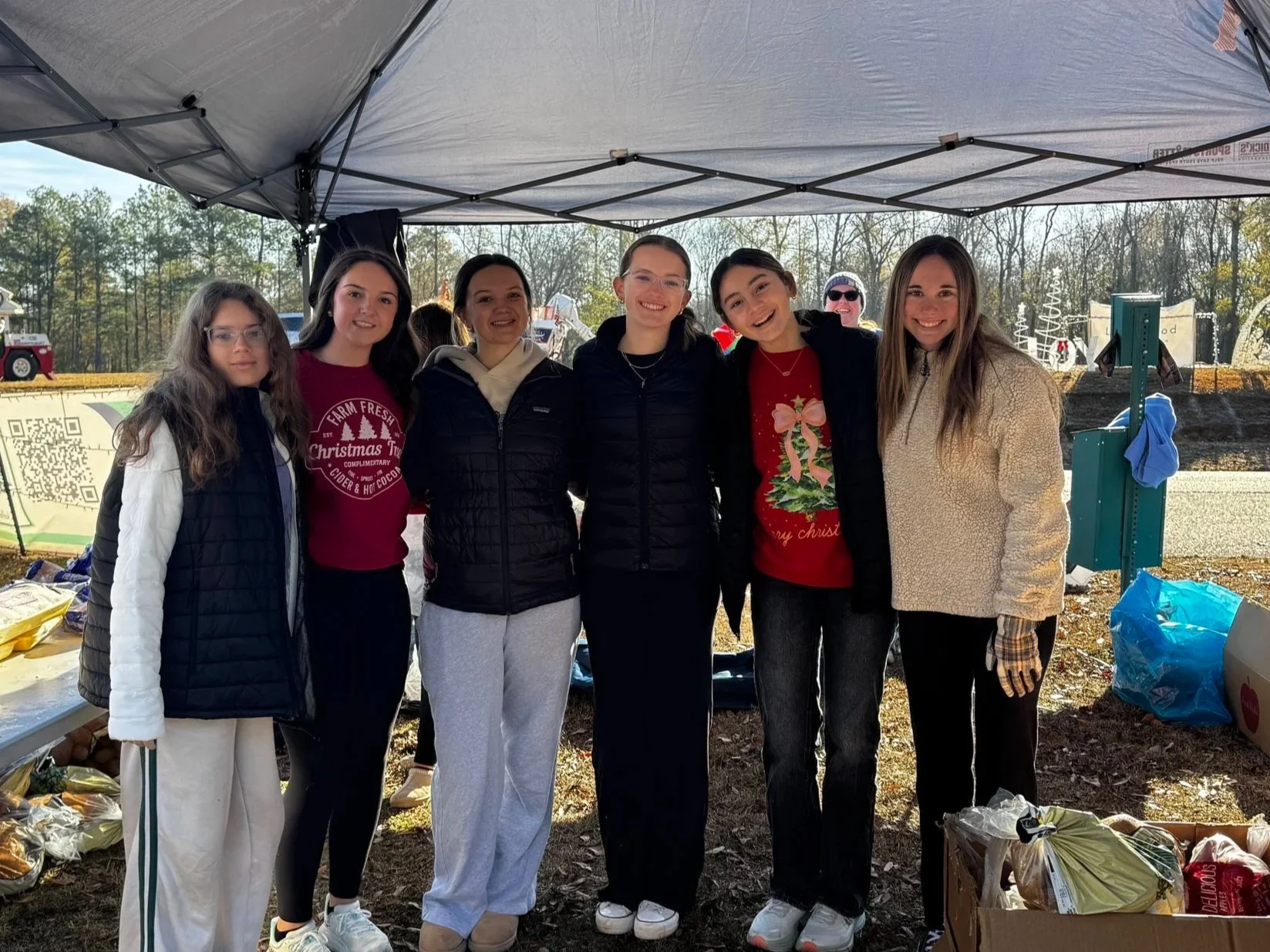 Group of seven young women standing under a canopy outdoors, smiling at camera, with trees and farm setup in background, during daytime.