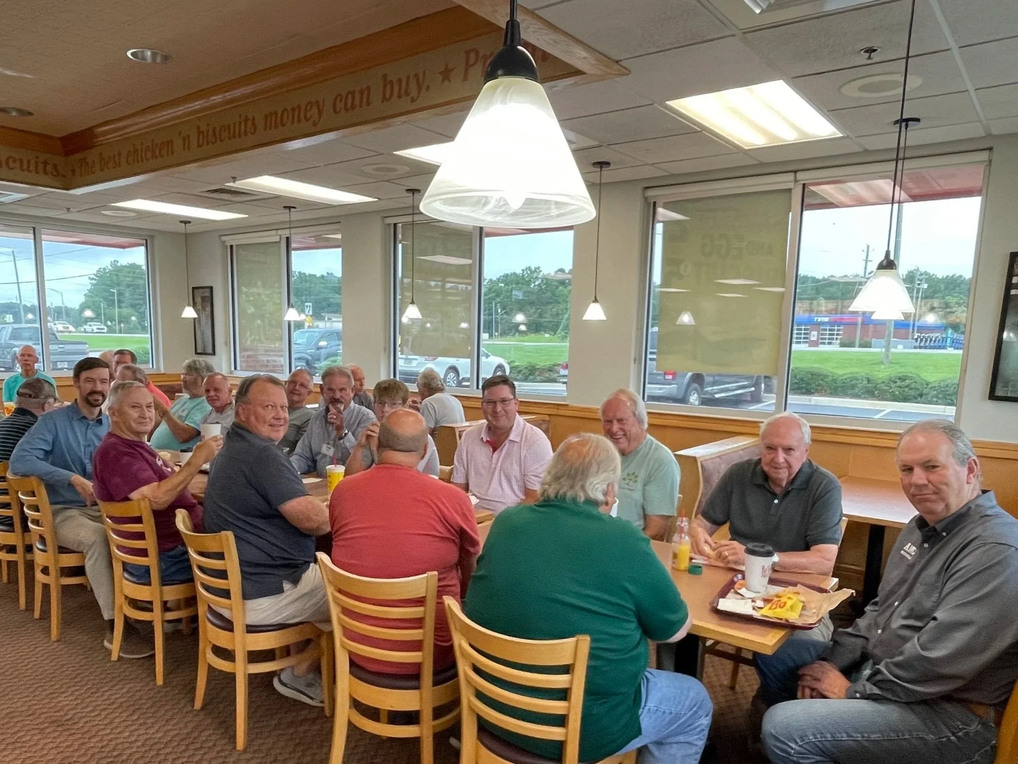 Group of people seated at a table inside a restaurant, smiling and enjoying drinks, with large windows showing parked cars outside.