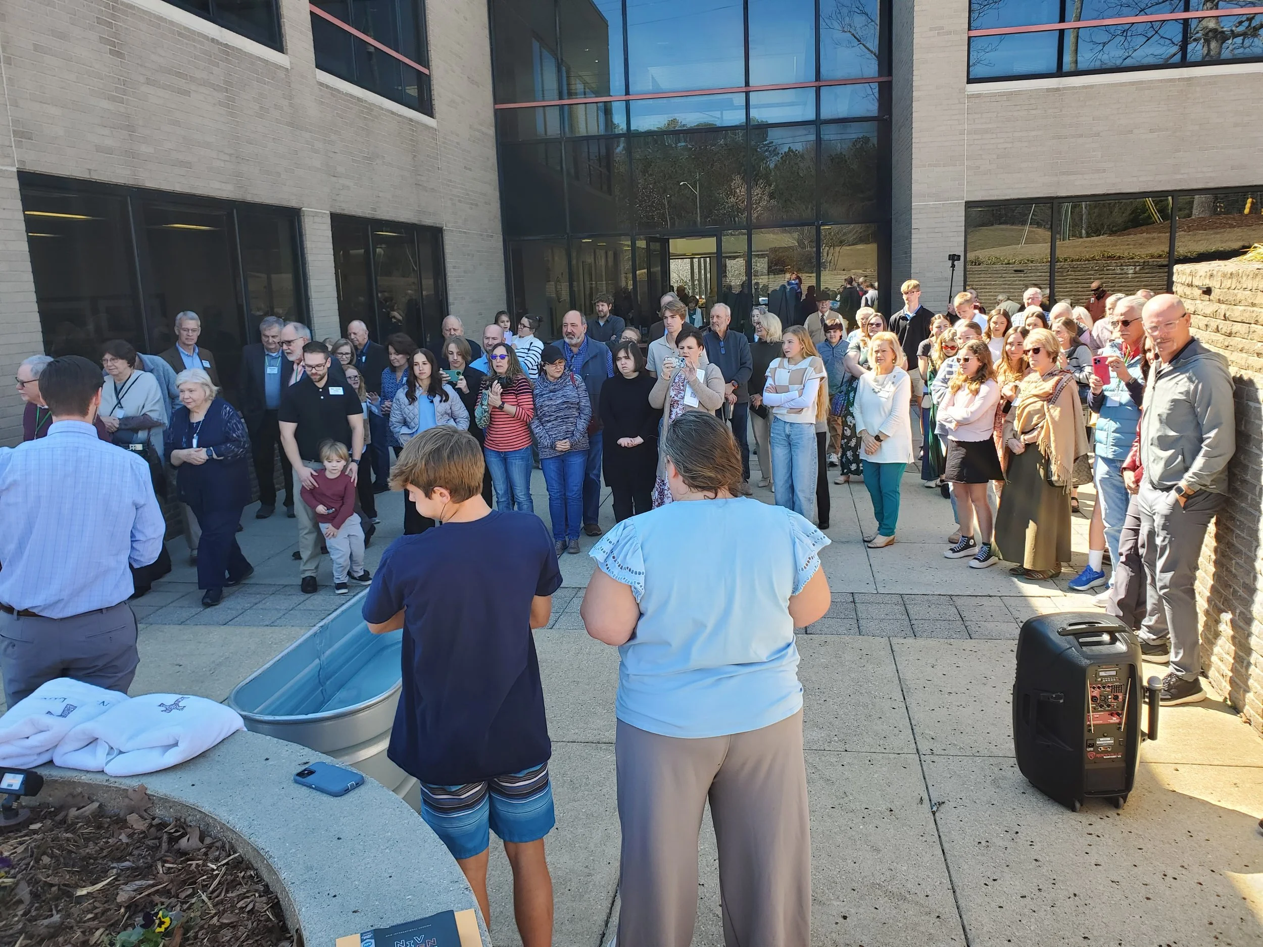 A large group of people gathered outside a modern building, attending an event or ceremony. Some are taking photos or videos, with a speaker or presenter visible at the front. The scene is well-lit with sunlight.