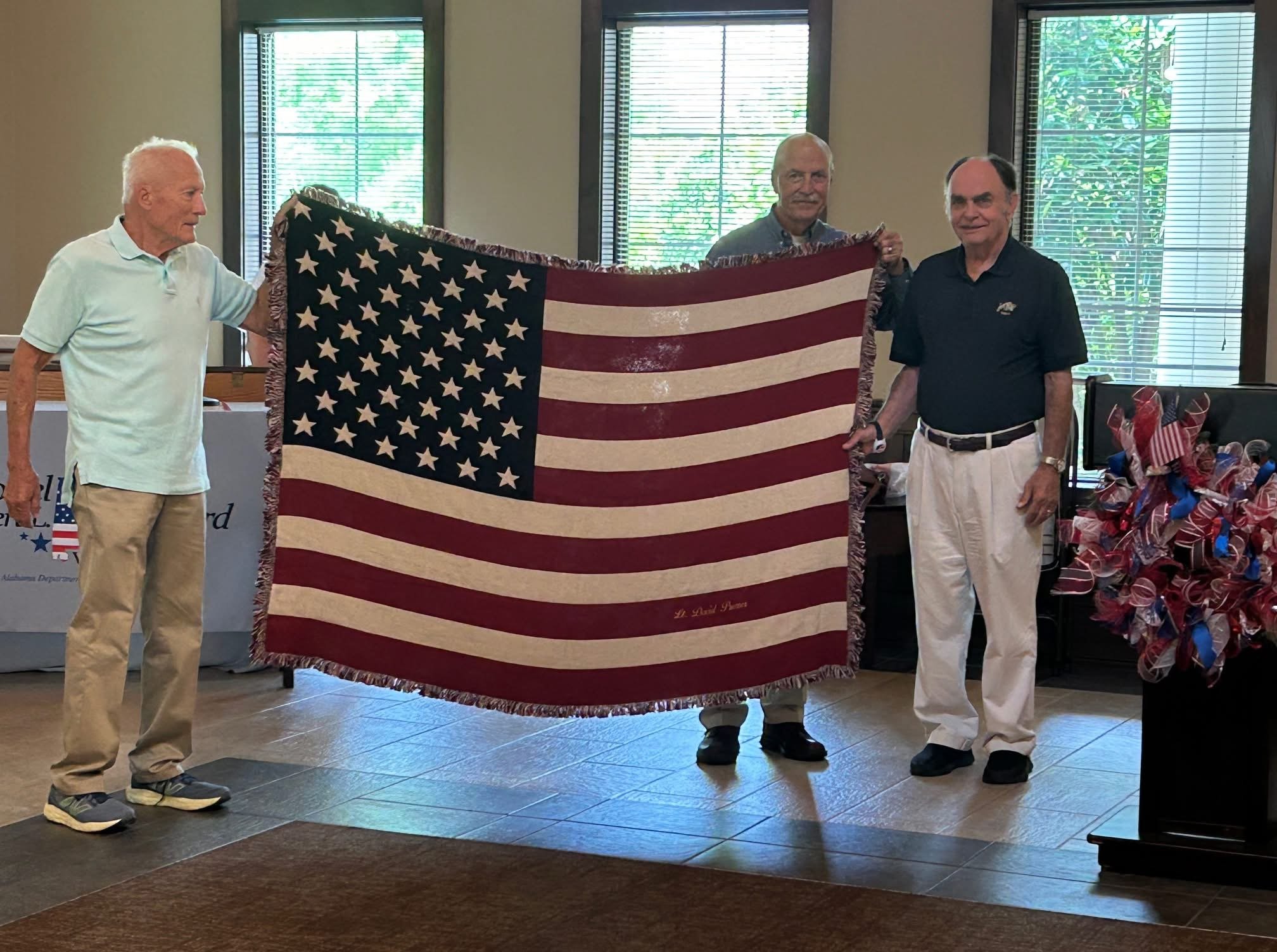 Three men holding a large American flag indoors with windows and patriotic decorations in the background.
