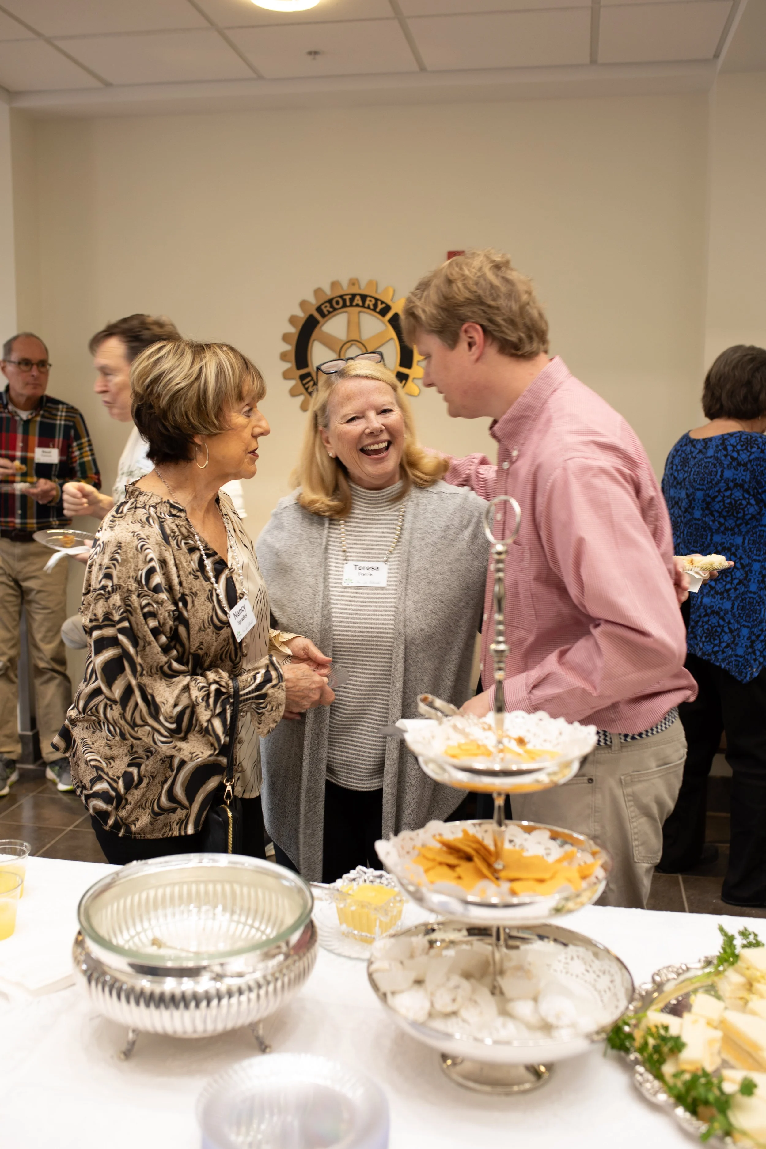 People socializing at a Rotary Club event, talking and smiling, with a table of desserts including cookies and chips in the foreground.